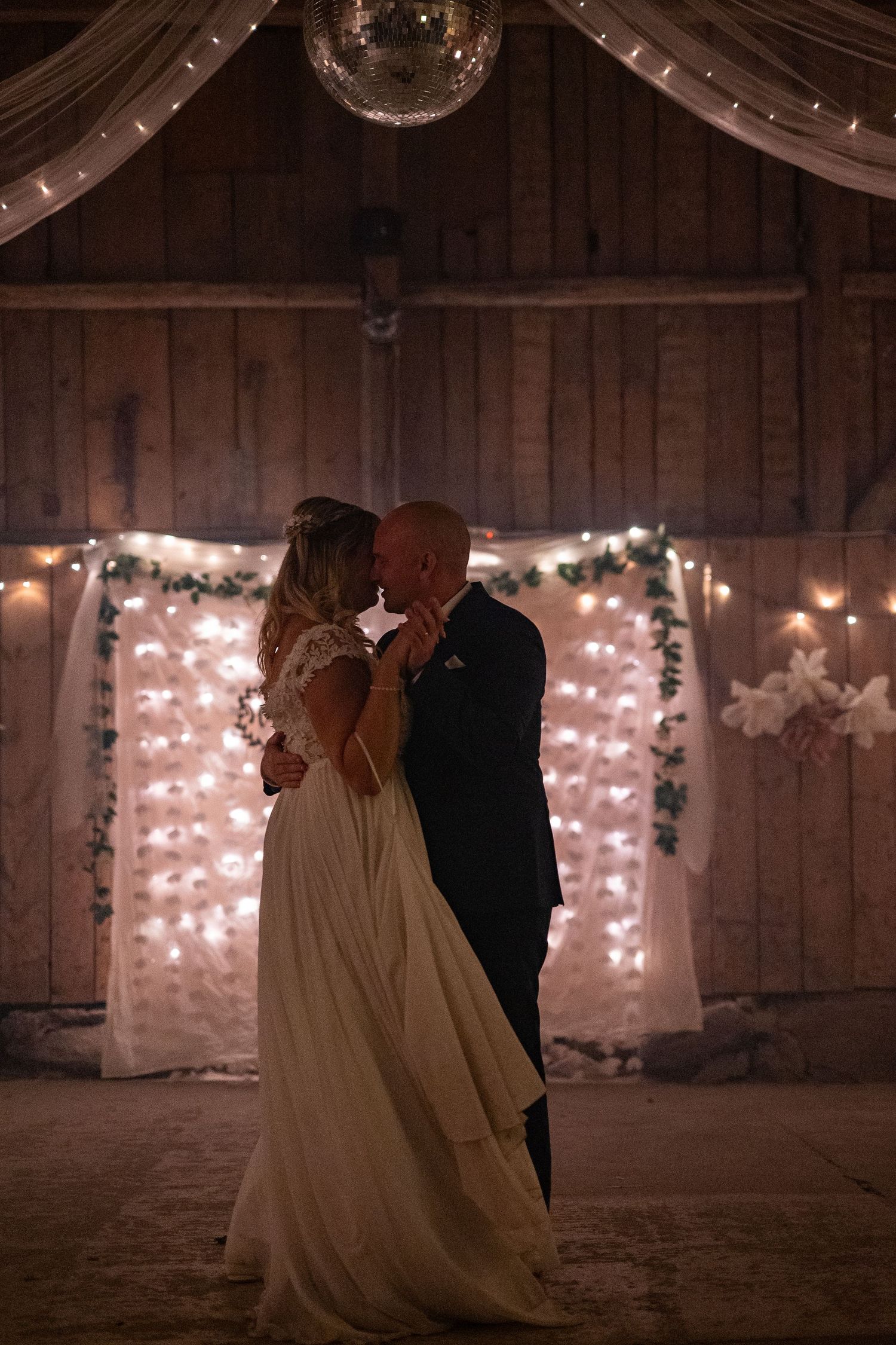 Bride and groom sharing first dance in a rustic barn wedding venue with fairy lights and floral backdrop.