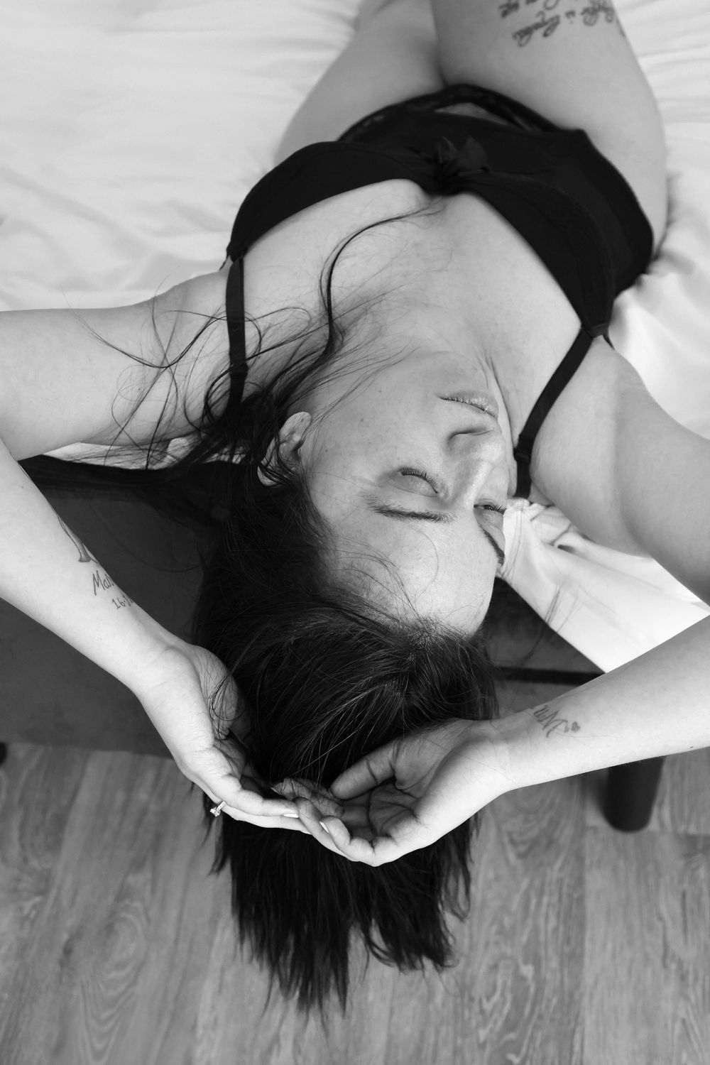 Black and white boudoir photo of a woman with dark hair lying back on a bed wearing black lingerie.