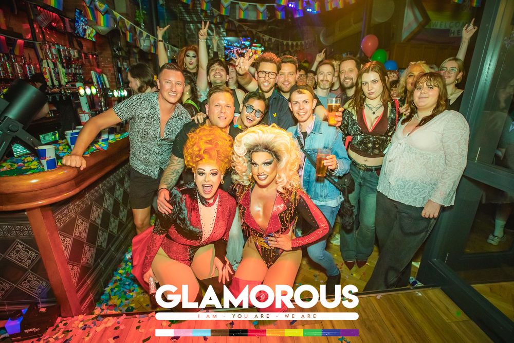 Glamorous drag queens pose with a lively crowd at a colorful nightclub event, wearing vibrant costumes and big hair.