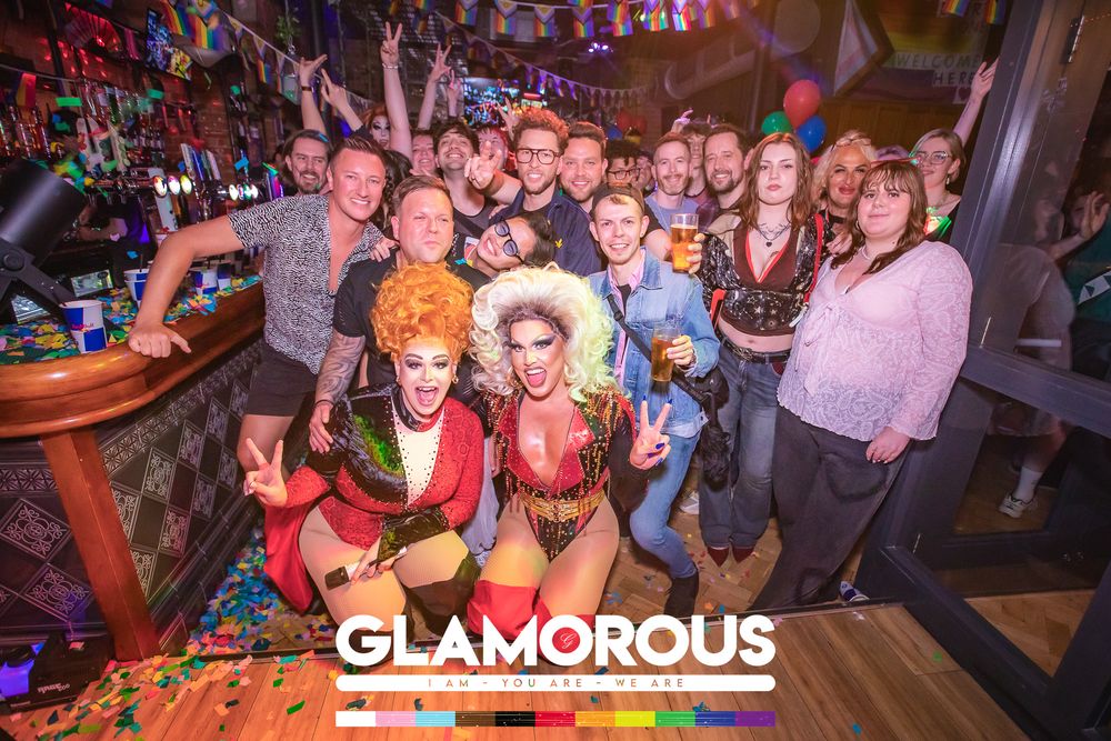 Glamorous nightclub event with drag queens and guests posing together at a vibrant, colorful bar celebration.