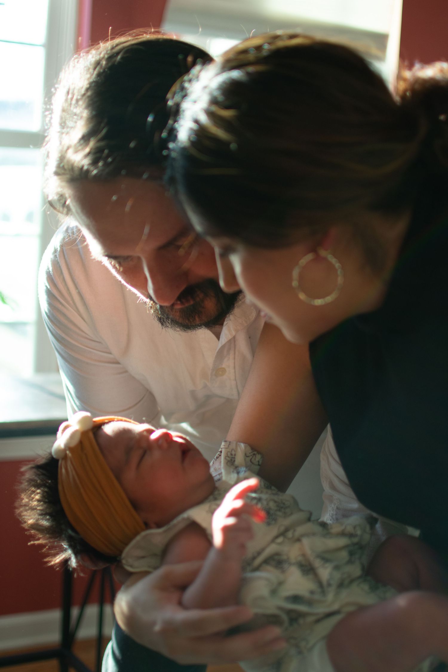 A couple lovingly gazes at their newborn baby wrapped in a blanket with a yellow bow headband near a sunlit window.