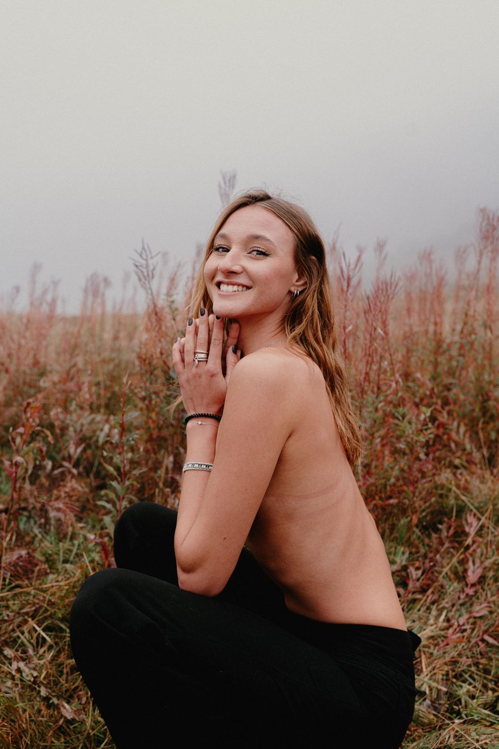 A smiling young woman sits in a field of tall autumn grasses on a misty day, wearing black pants and silver jewelry.
