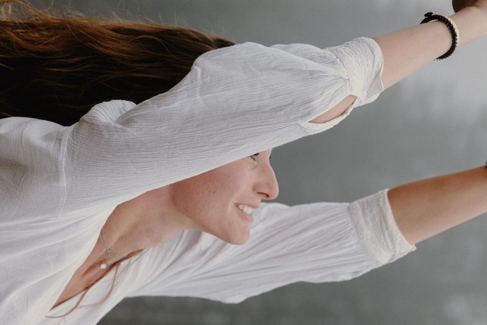 A smiling woman in a white linen shirt with arms outstretched, hair flowing in the wind against a gray background.