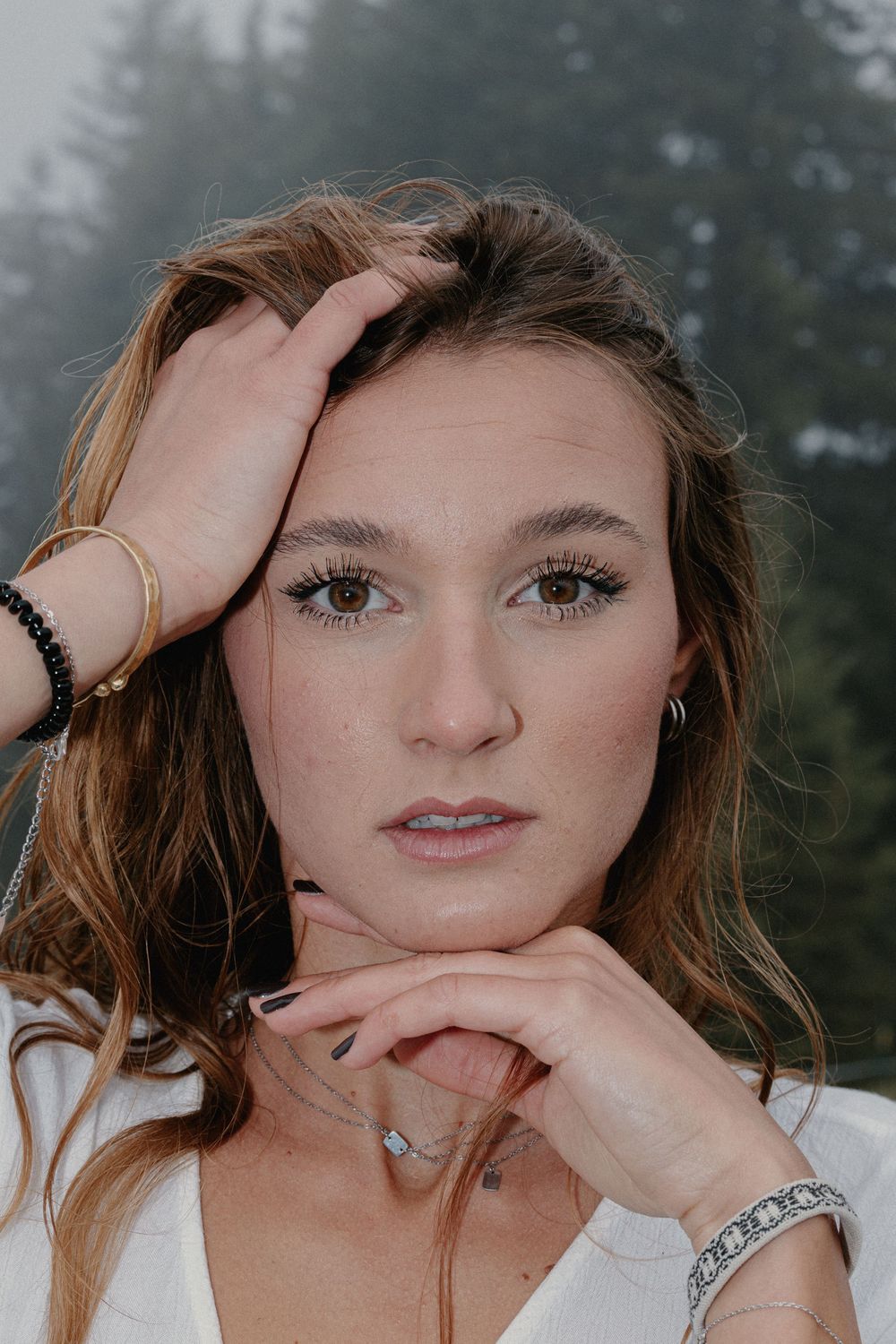 Young woman with brown eyes and freckles posing with hands near face, wearing gold and silver bracelets.