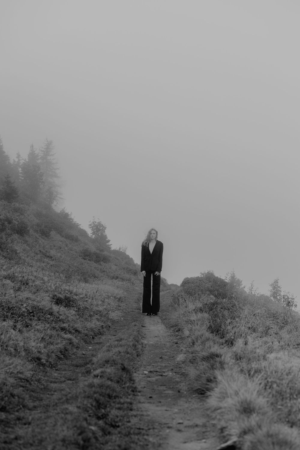 Person standing alone on a misty mountain trail surrounded by fog and wild vegetation in black and white.