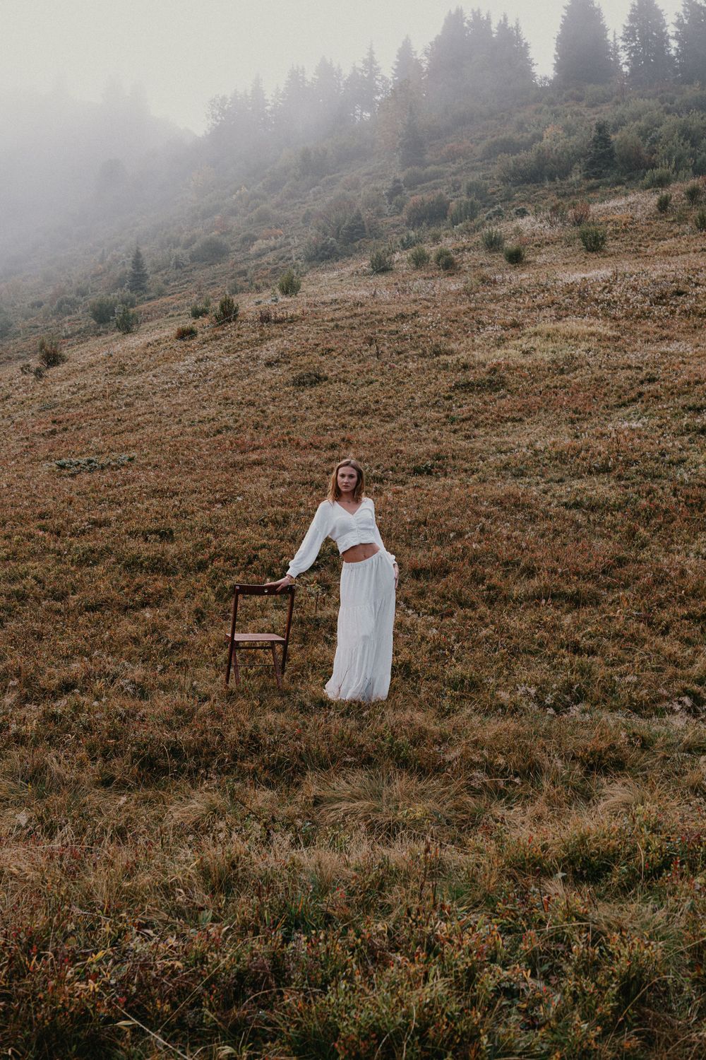 Woman in white outfit standing beside a wooden chair on a misty mountain meadow with foggy trees in background.