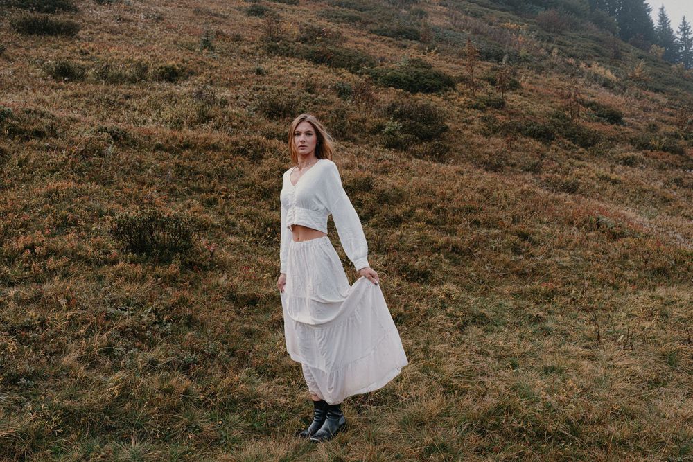 Woman in white crop top and layered midi skirt stands on a grassy hillside in a serene outdoor setting.