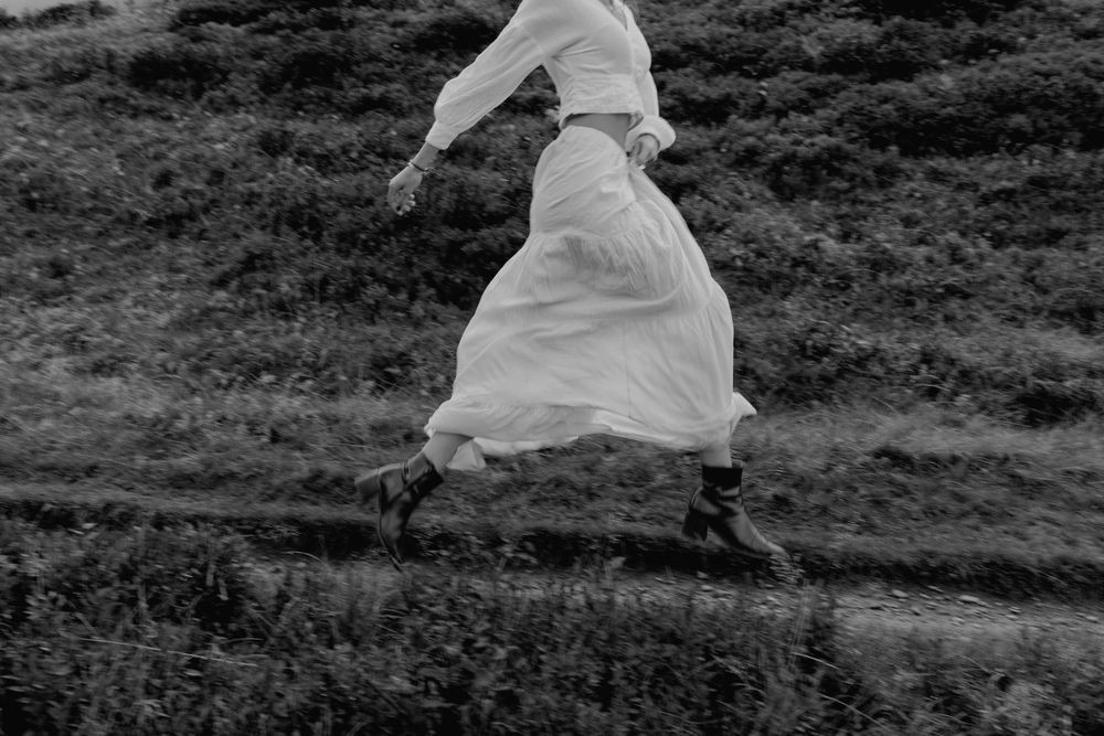 Black and white photo of a woman in a white dress and boots running through a grassy field.