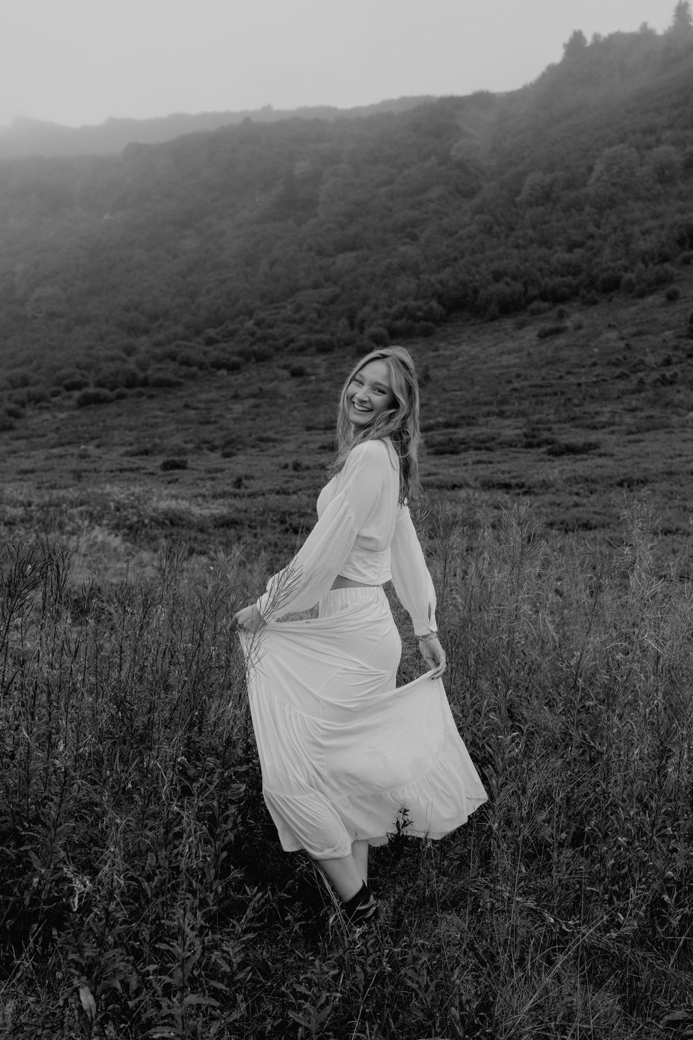 Smiling woman in white dress twirling in grassy mountain meadow, elegant black and white photo.