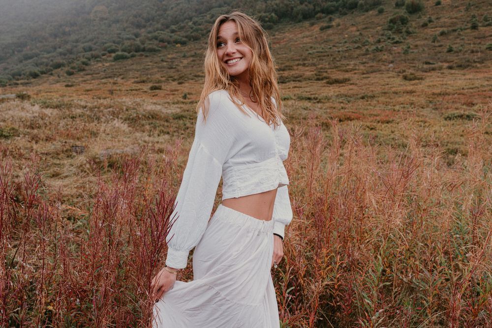 Woman in white crop top and skirt smiling in a scenic field with red grasses and misty hills.
