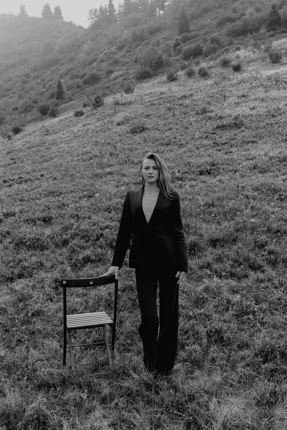 Black and white photo of a woman in a dark suit standing on a grassy hillside next to a wooden chair.