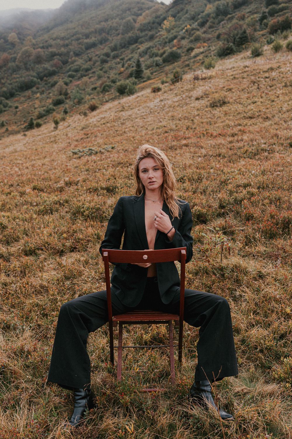 Woman in dark blazer and trousers sits on wooden chair in autumn mountain meadow landscape.
