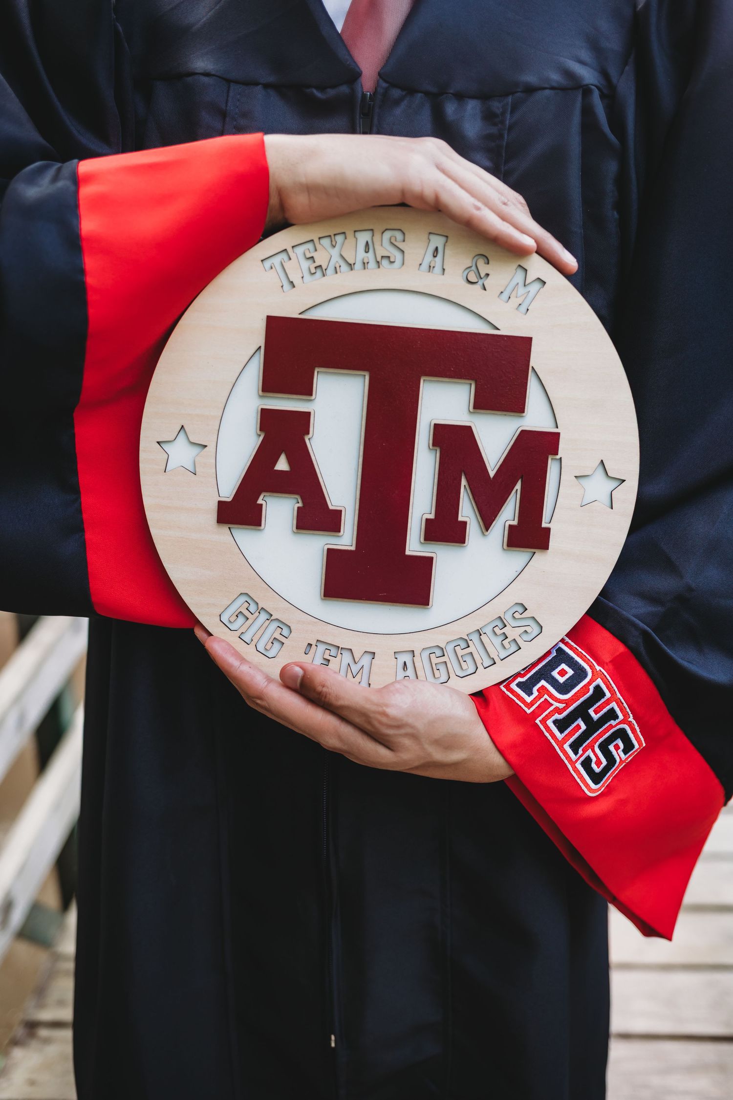 Graduate in black gown holds wooden Texas A&M ATM logo sign with Gig Em Aggies text and red sash.
