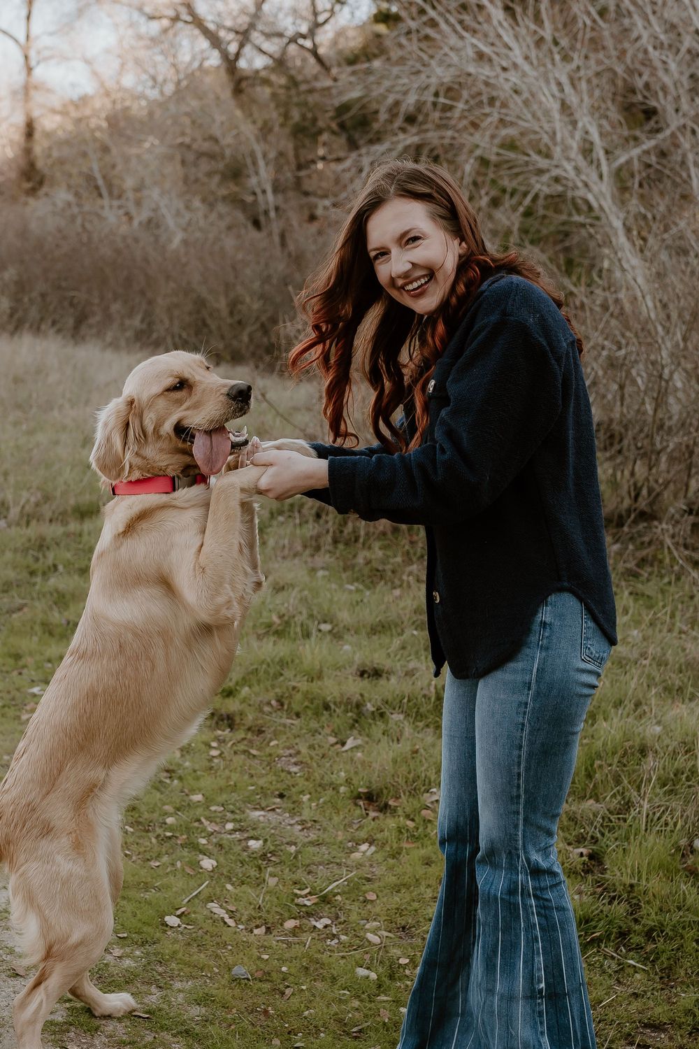 A smiling woman with red hair plays with her golden retriever outdoors, holding its paws as it stands up.