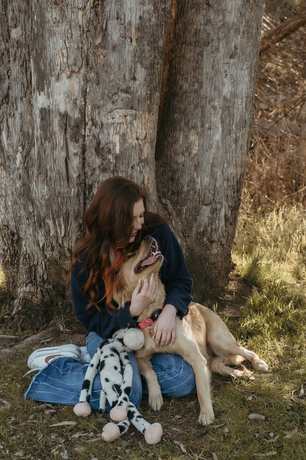 Young woman with red hair sitting against a tree trunk, cuddling a golden dog with a dalmatian toy outdoors.