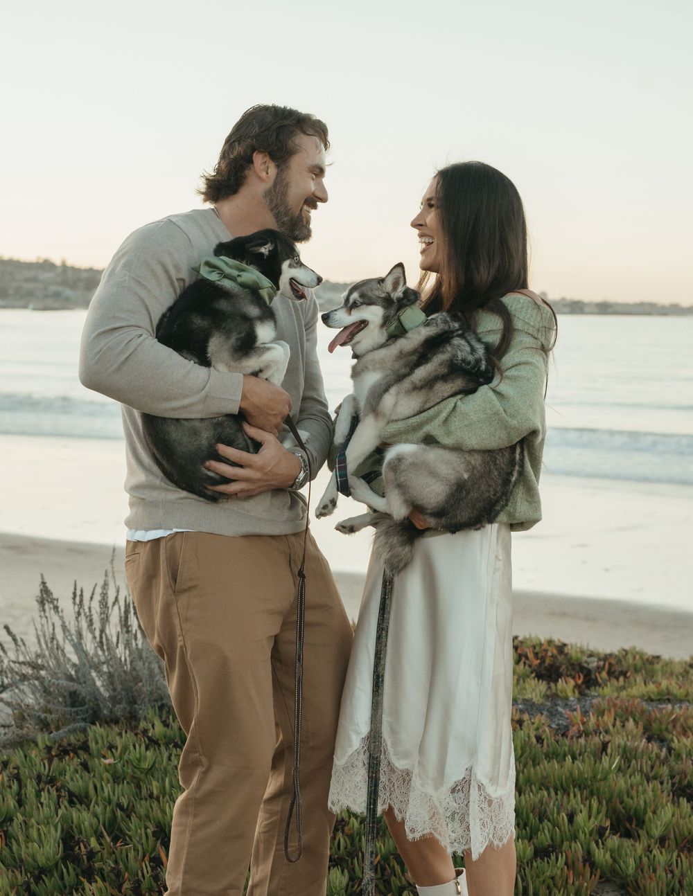 Happy couple holding their Husky dogs on a scenic beach at sunset, smiling at each other near the ocean.