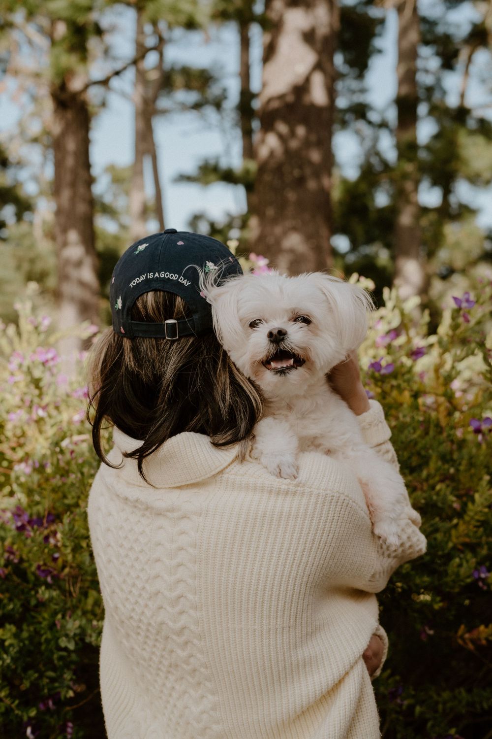 Woman in baseball cap holds fluffy white Maltese dog outdoors among colorful wildflowers and trees.