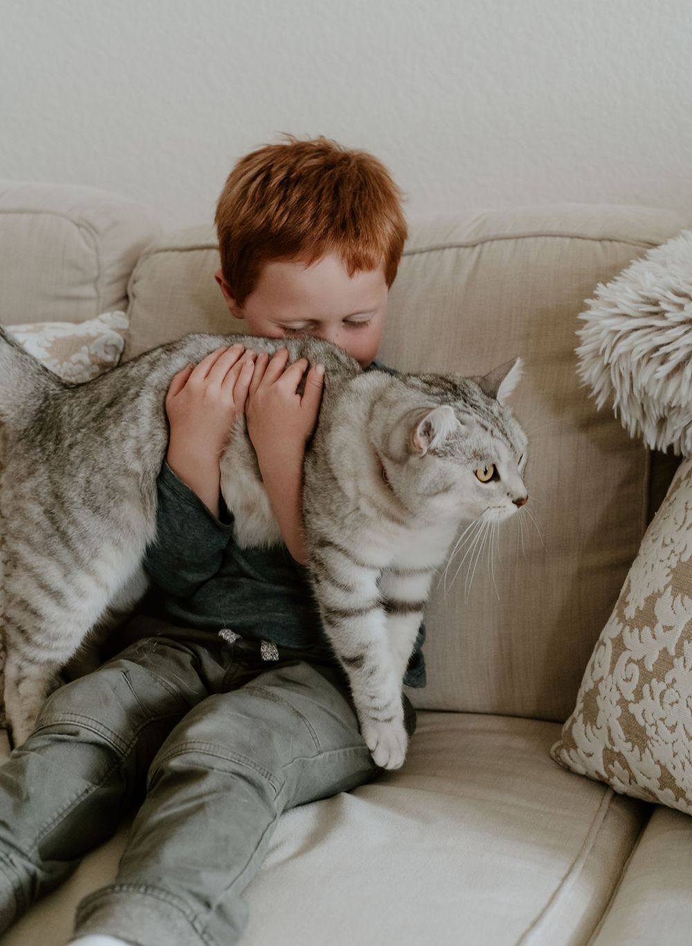 A red-haired boy hugging a silver tabby cat on a beige sofa with decorative pillows.