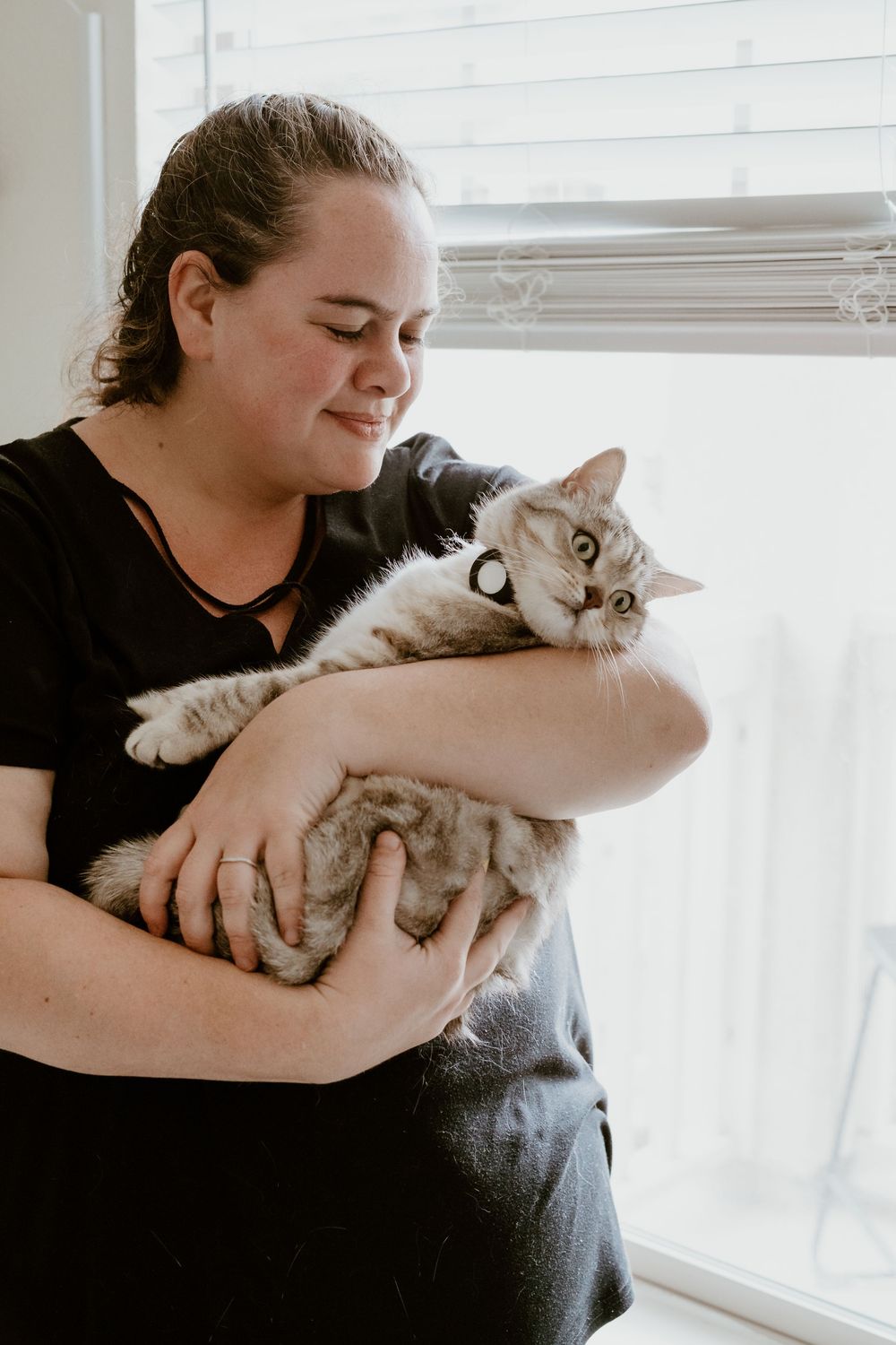 Woman in black shirt smiling while holding a fluffy gray tabby cat near a bright window indoors.