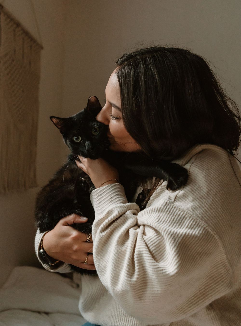 A woman in a cream sweater lovingly kisses her black cat while holding it gently in her arms indoors.