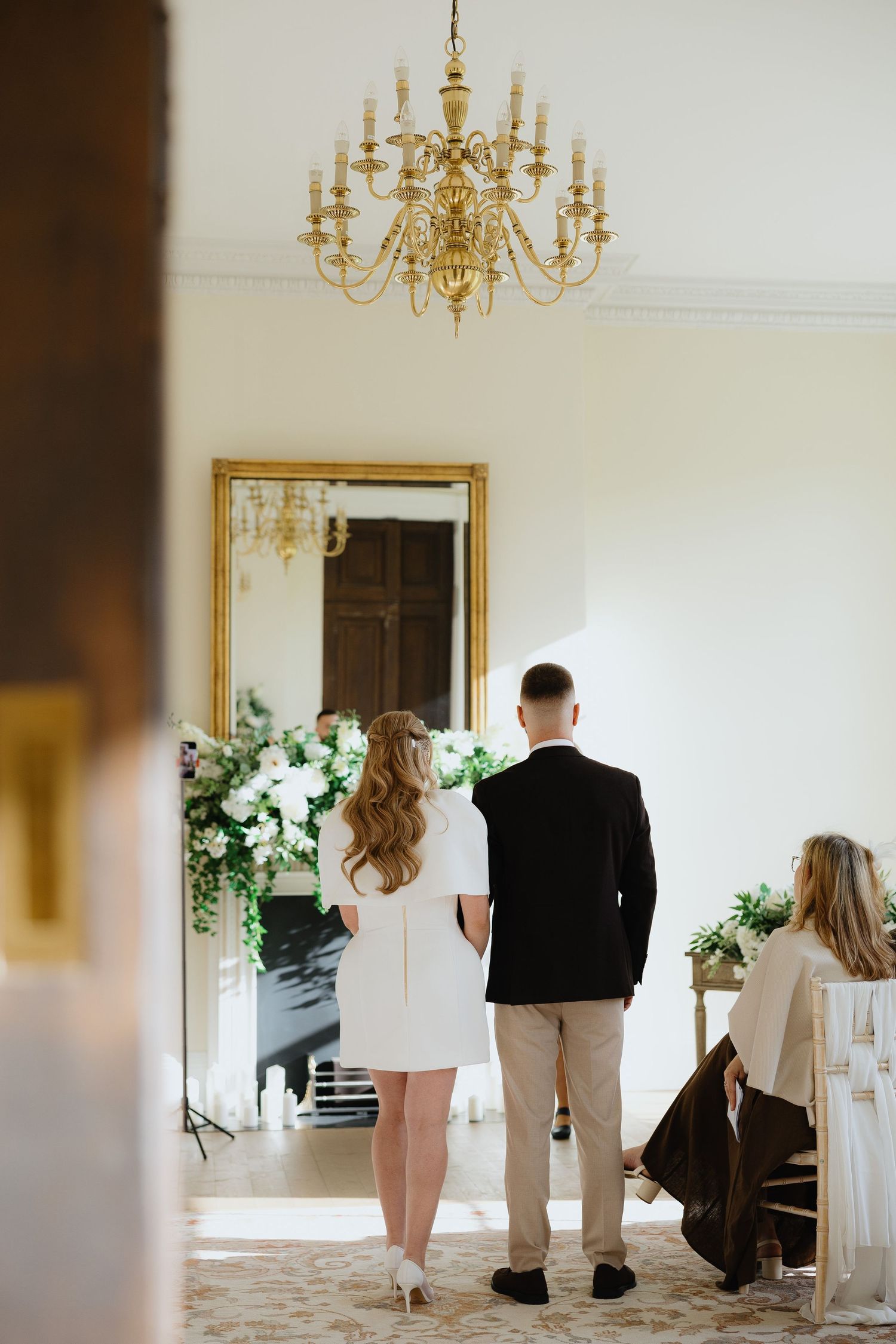 Couple standing before a grand mirror with floral arrangements under a gold chandelier in an elegant white room.