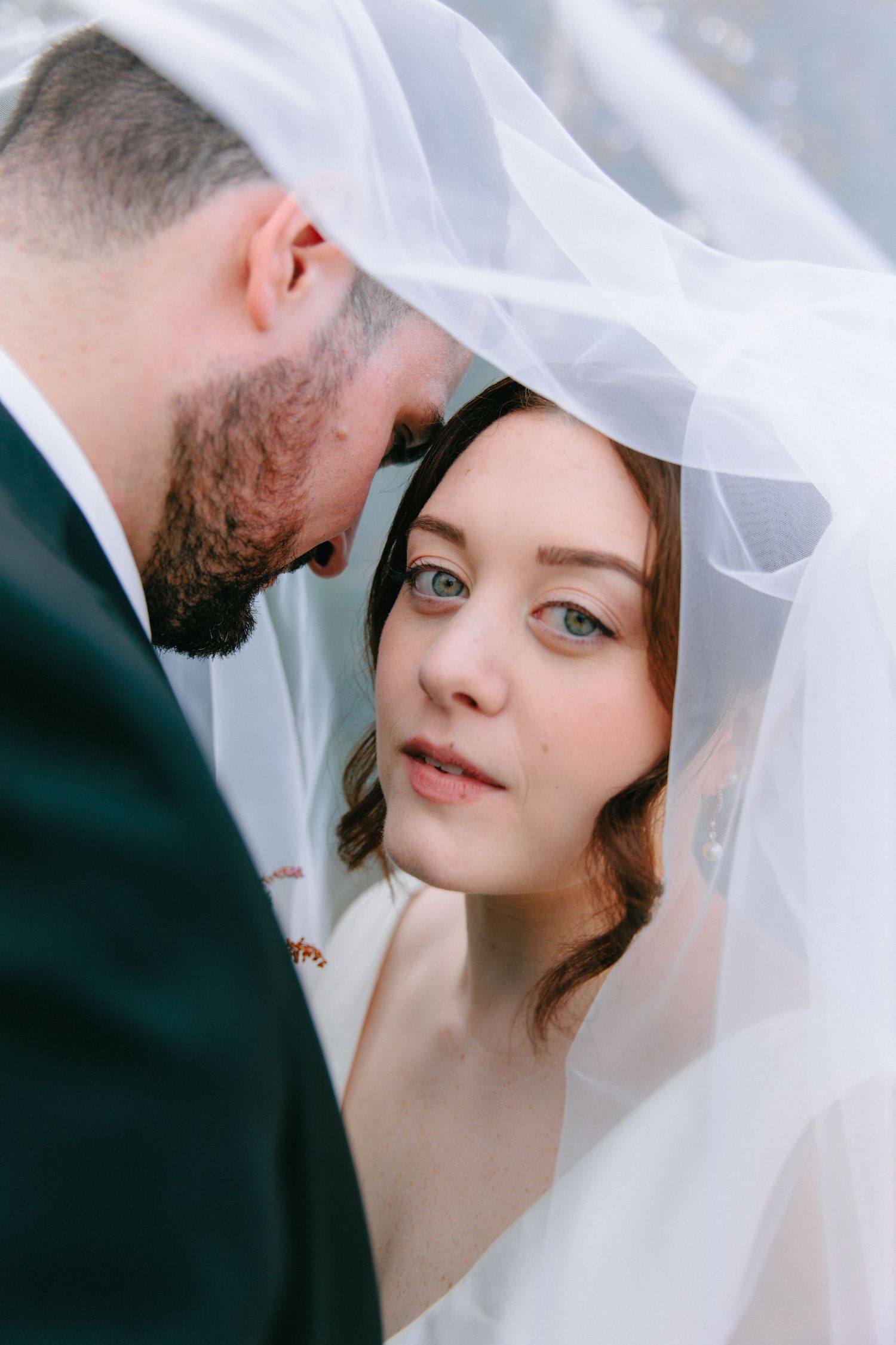 Bride and groom share an intimate moment under a flowing white veil, bride gazing up with blue eyes.