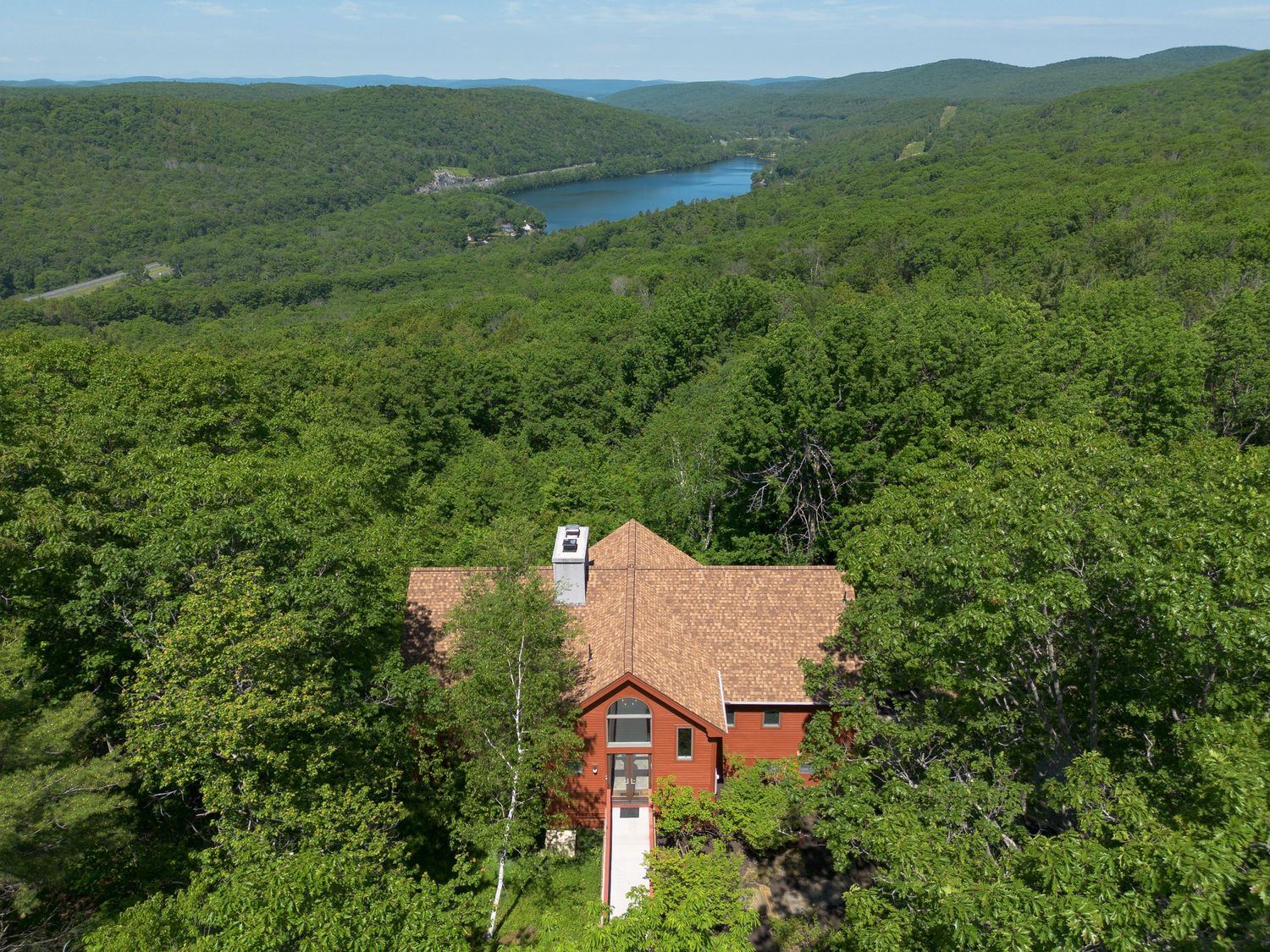 Aerial view of red home surrounded by dense forest with scenic lake backdrop.