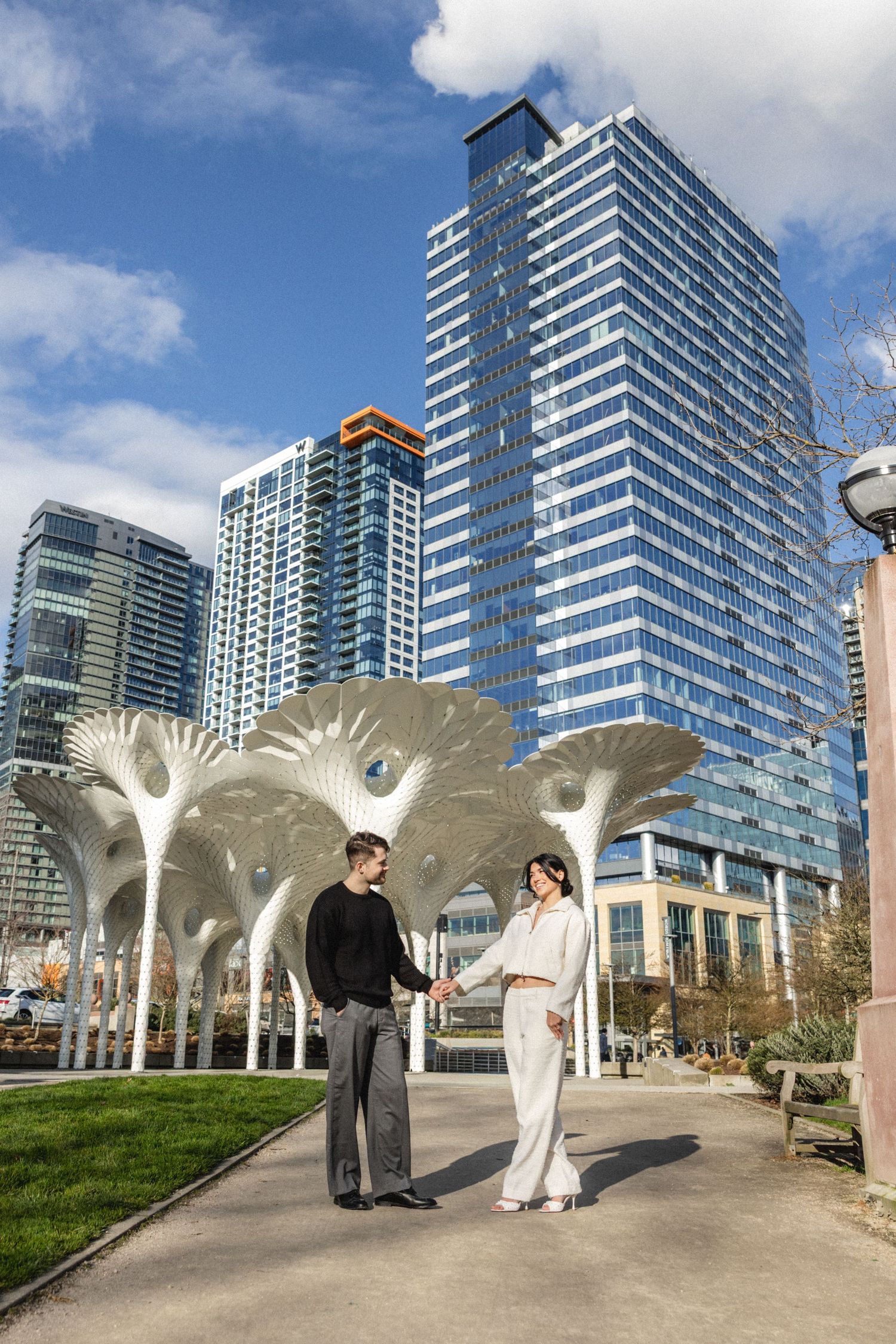 Couple holding hands in urban park with white sculptural canopy structures and modern glass skyscrapers under blue sky.