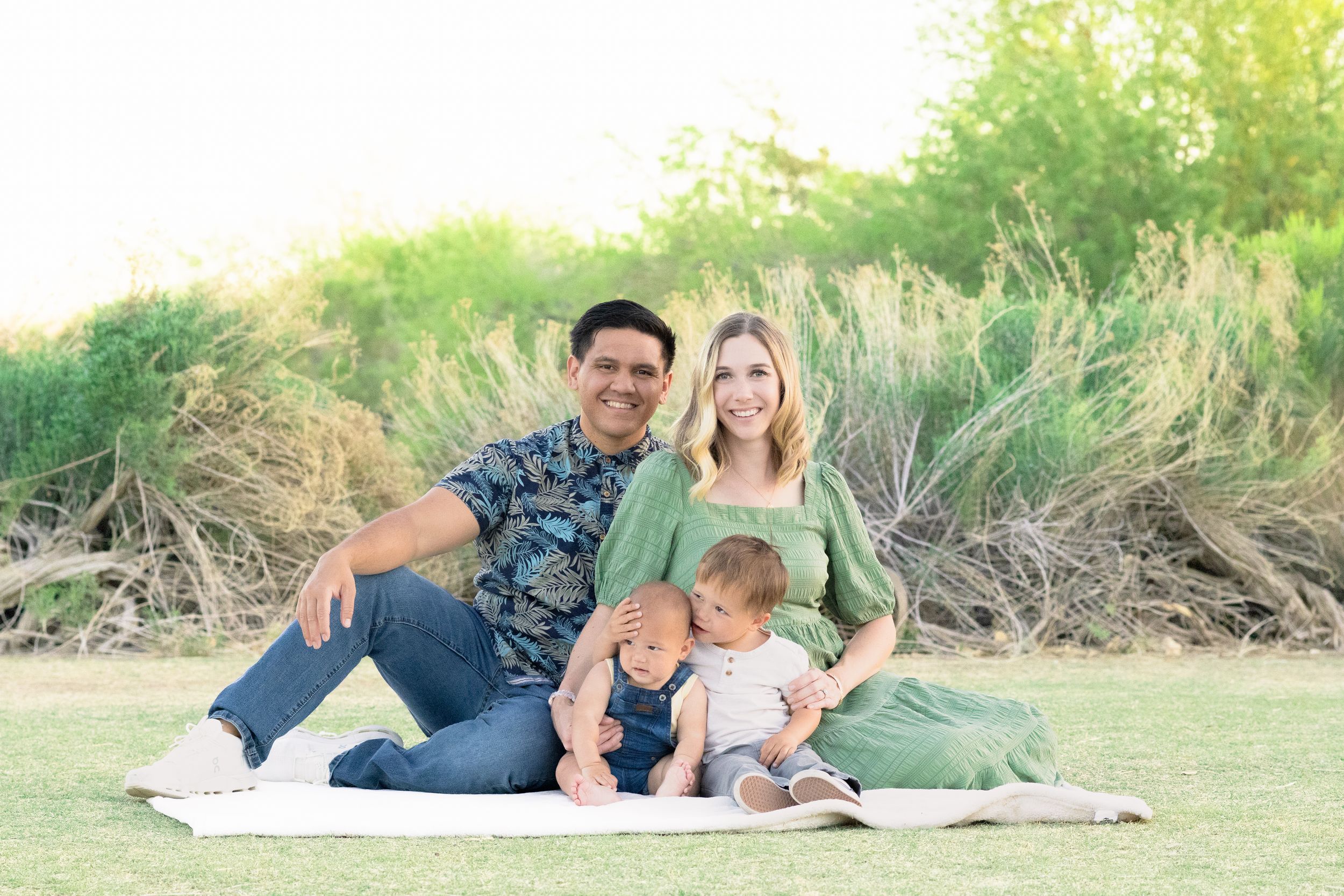 Young family of 4 sitting on a picnic blanket in front of greenery brush at the Riparian Preserve in Gilbert, Arizona.