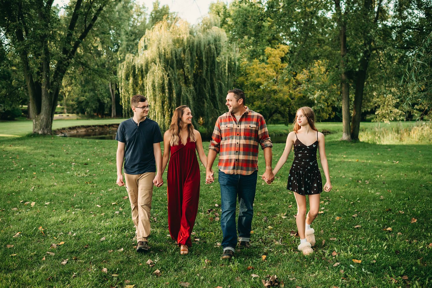 Four people walk hand-in-hand through a lush green park surrounded by willow trees during a family photo session.