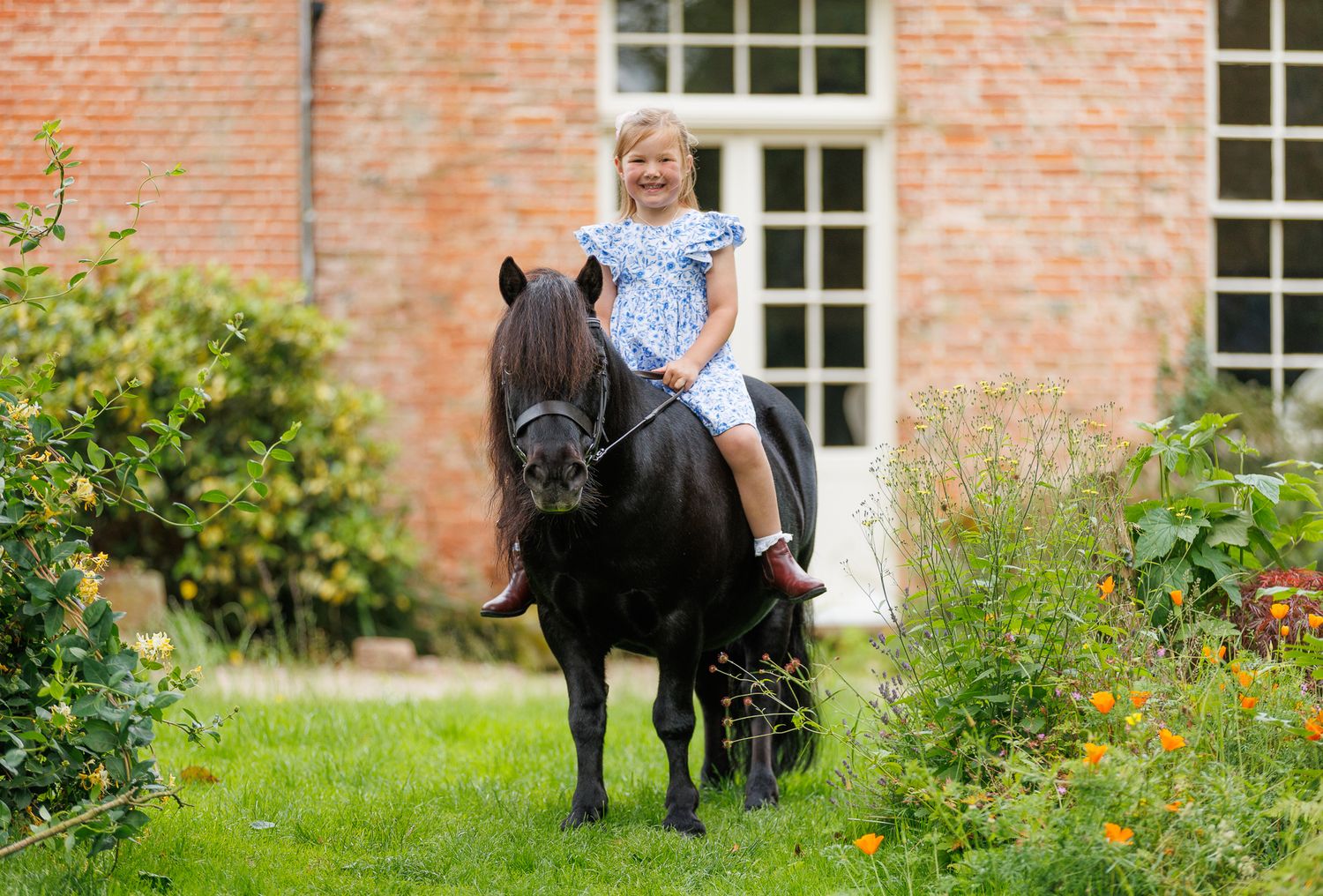 Woman leading a dark brown horse along a lush green garden path beside a tranquil pond.