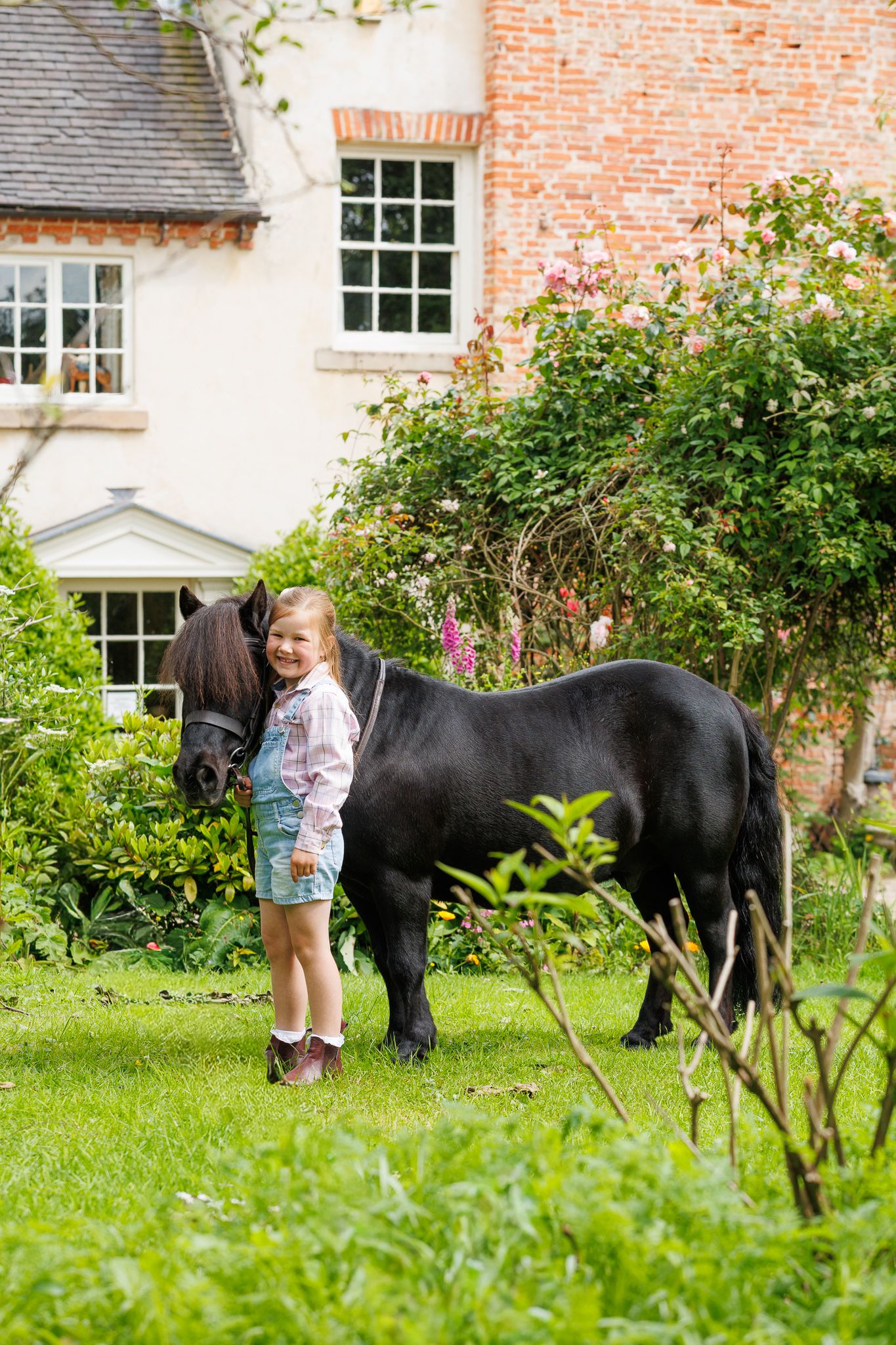 Smiling young girl in white dress crouching beside a black pony among daisies in a lush green garden.