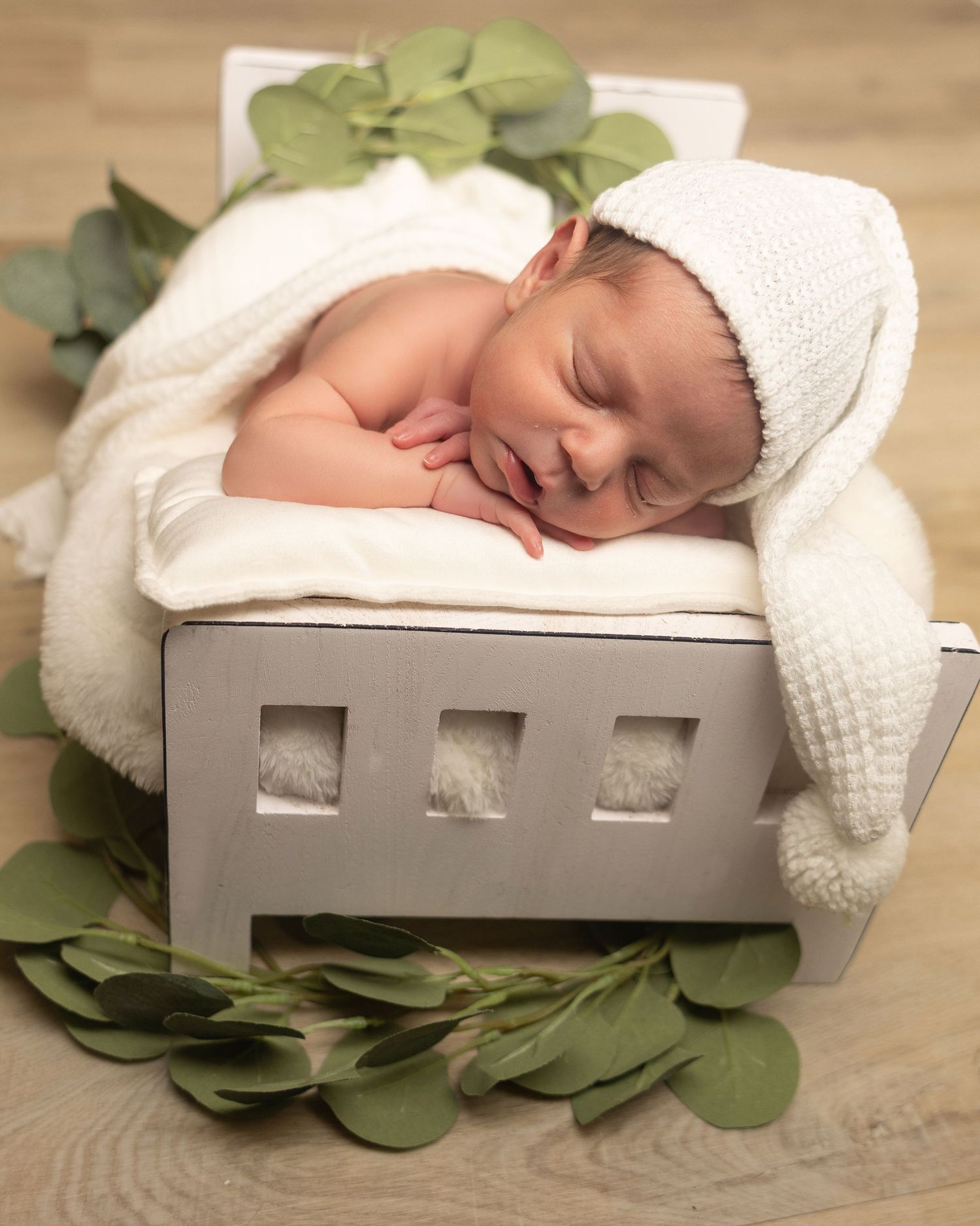 Sleeping newborn baby in white knit hat on miniature bed with green leaves.