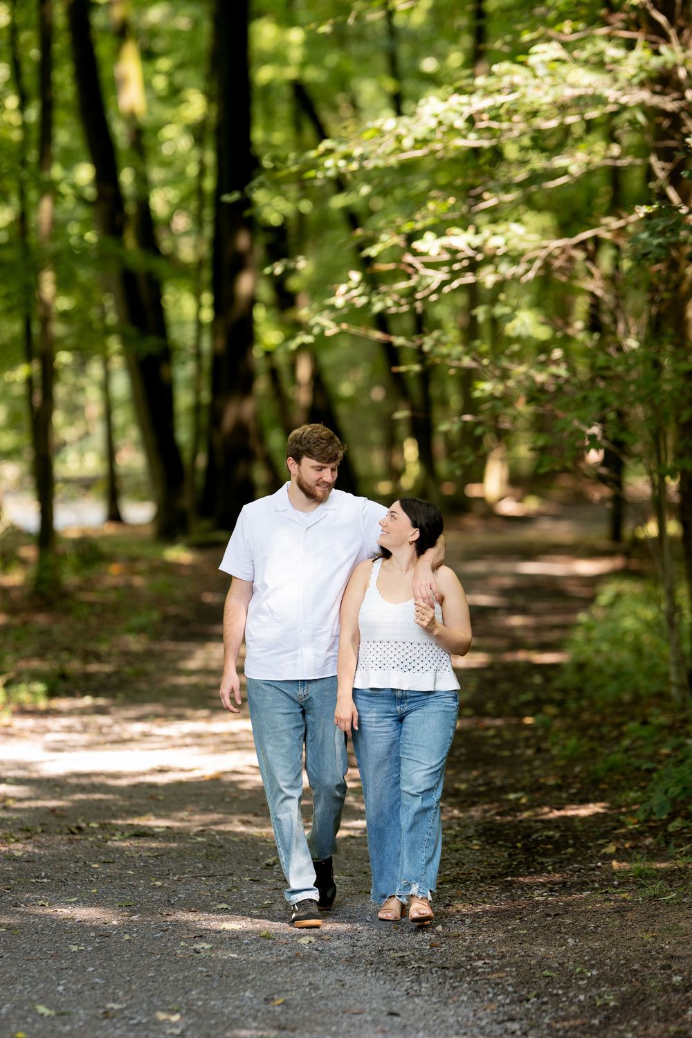 A happy couple walks hand-in-hand along a sunlit forest path, smiling at each other in casual attire.
