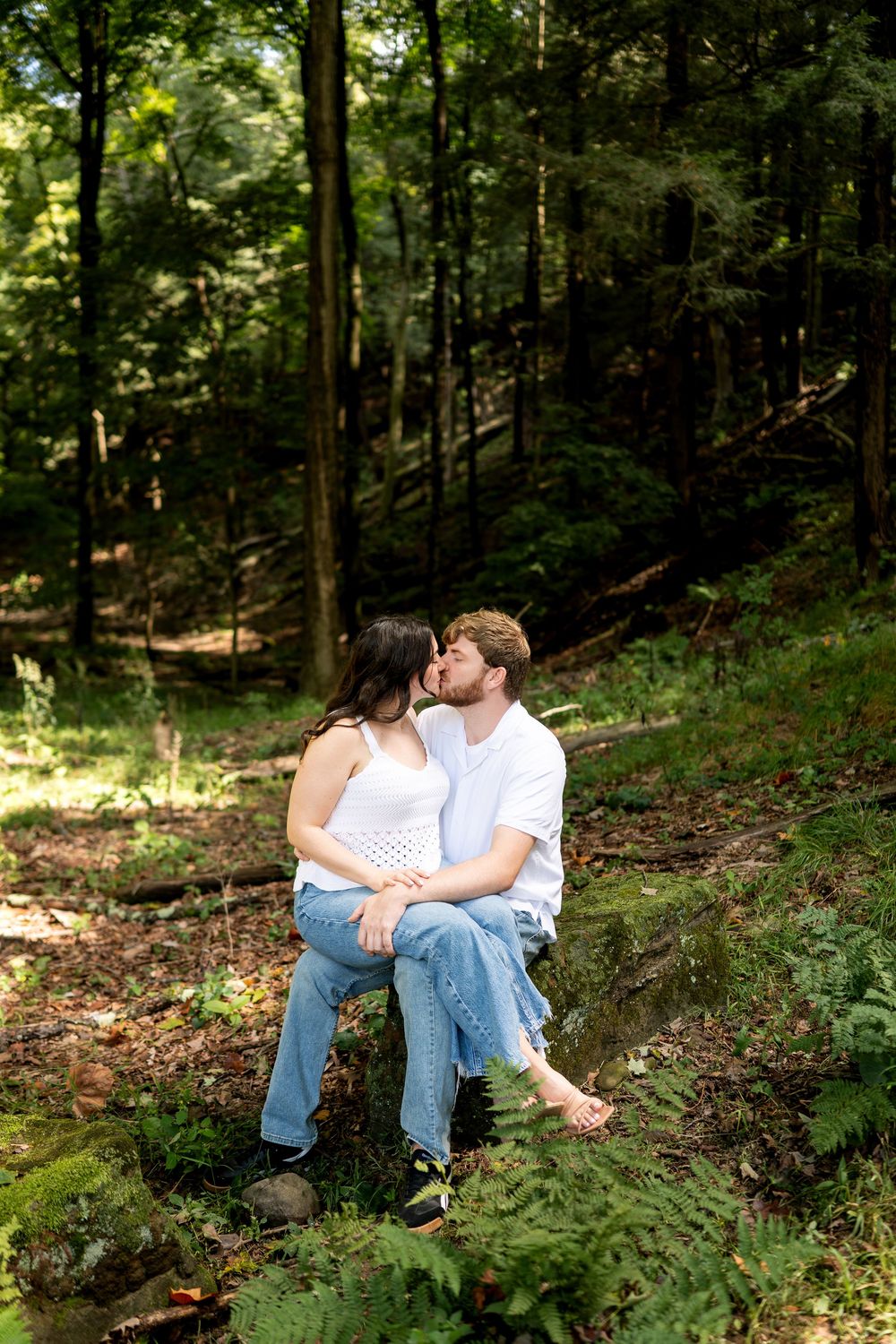 Couple sharing a kiss while sitting on rocks in a lush green forest setting.