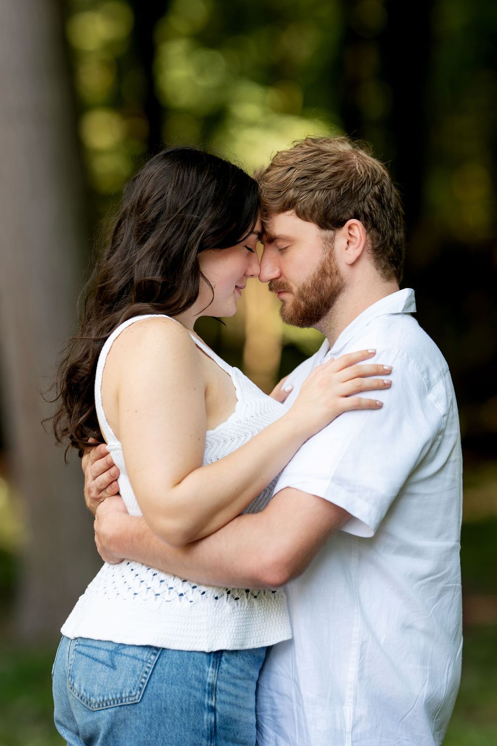 Young couple touching foreheads intimately while embracing in sun-dappled forest setting.