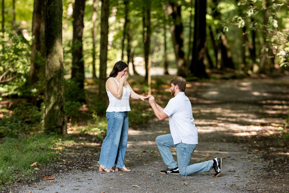Man kneeling on forest path proposing to surprised woman covering her face with hands in sunlit woods.