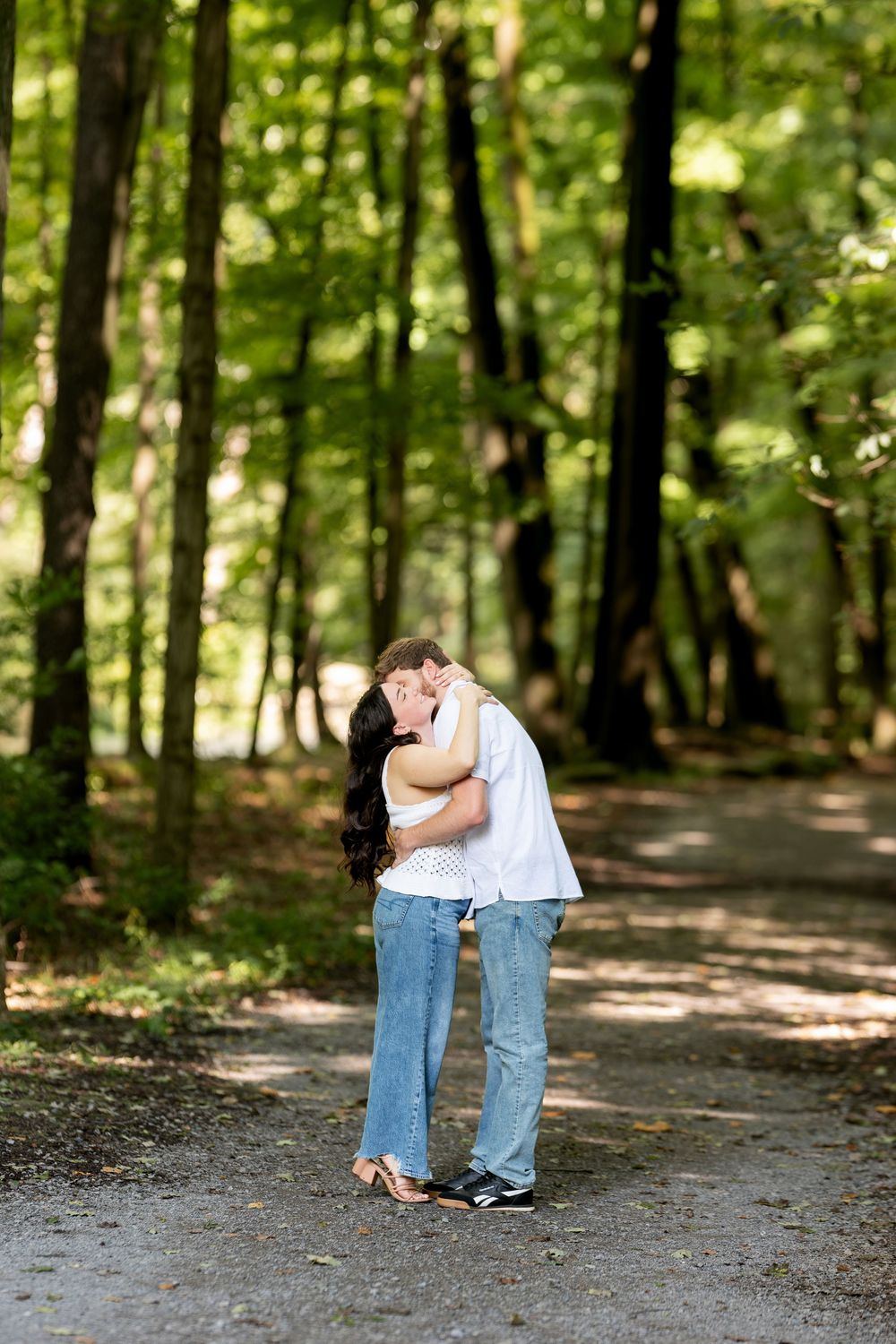 Couple sharing a kiss on a shaded forest path surrounded by tall green trees.