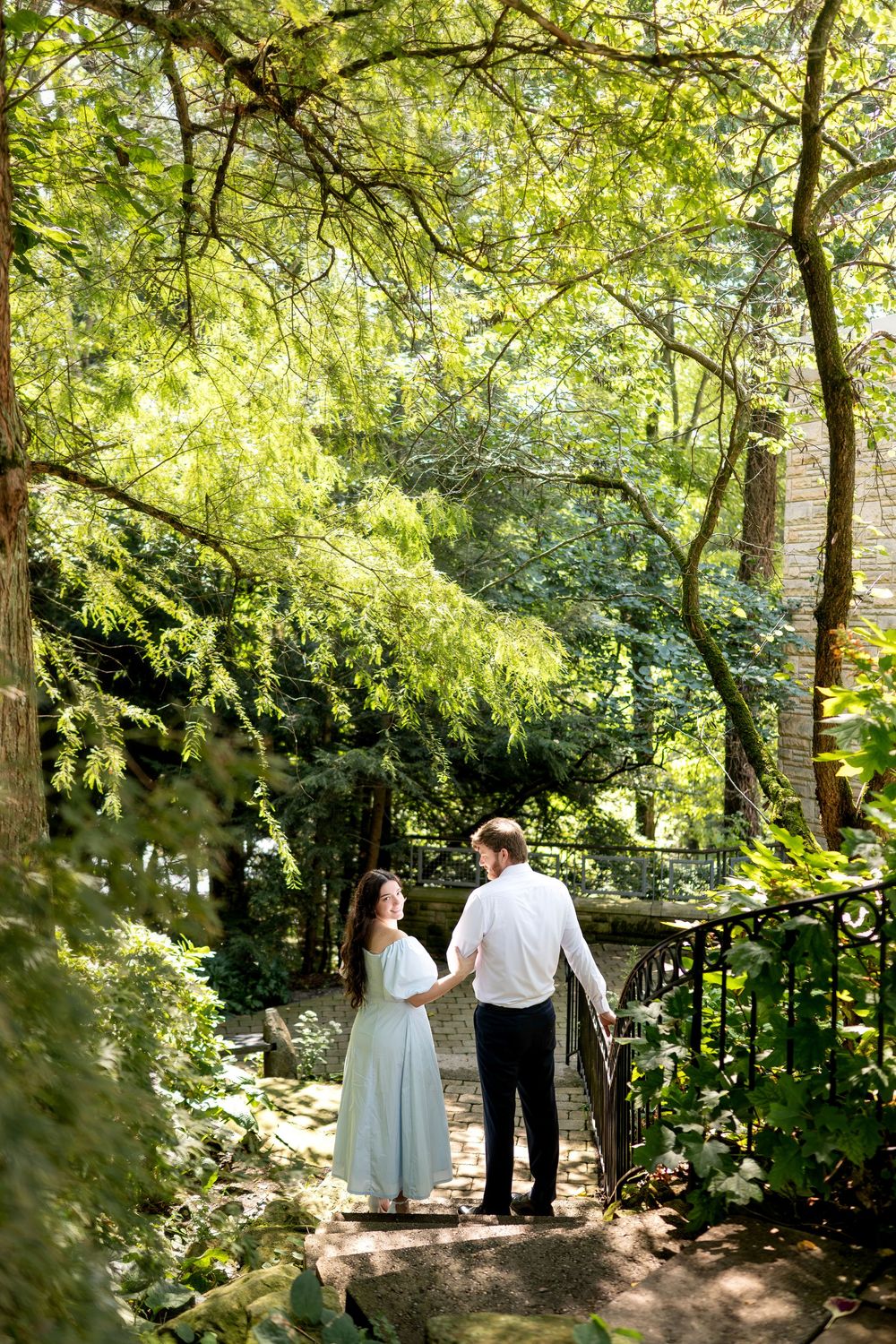 Couple holding hands on a sunlit garden path with lush green trees and iron railing.