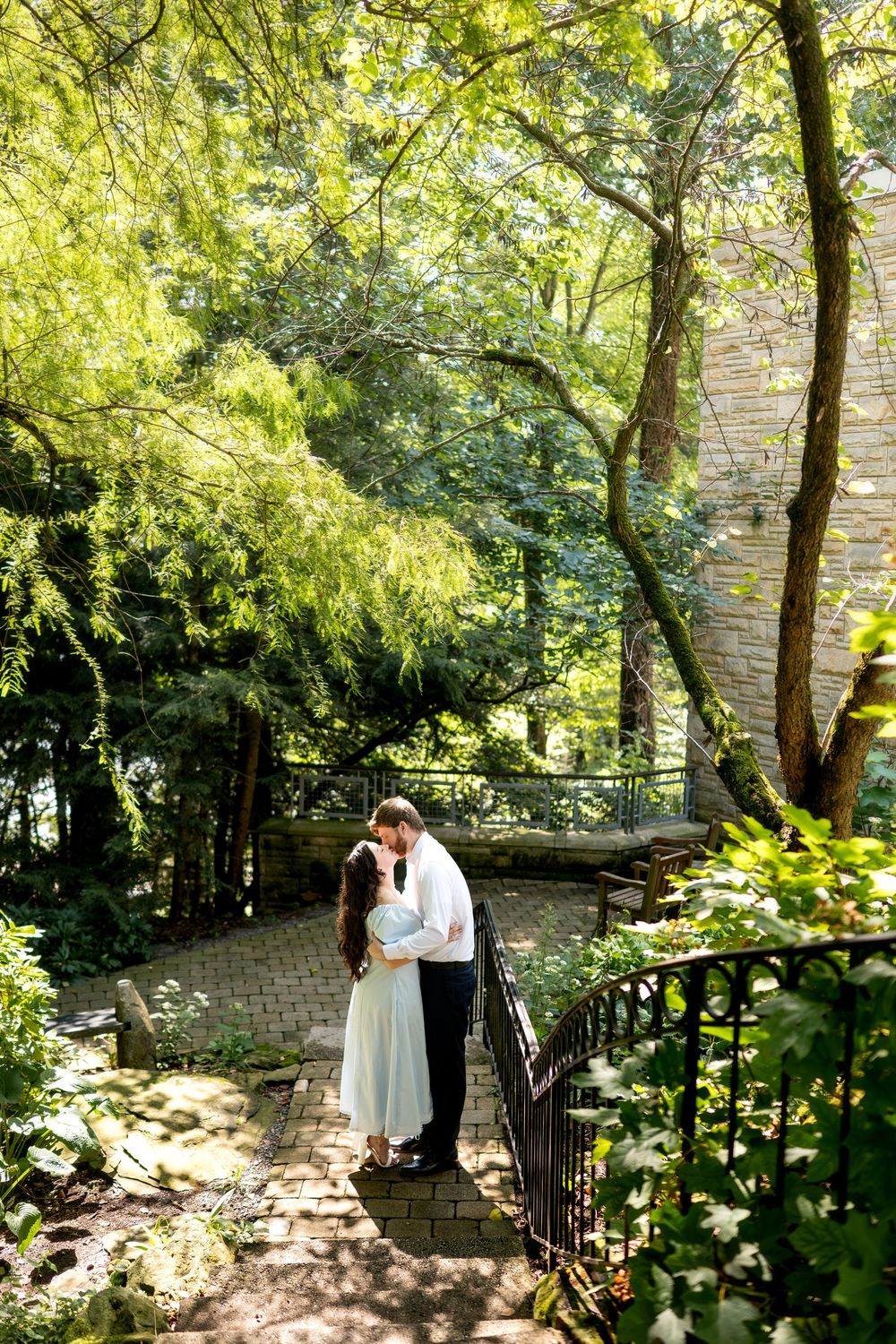 Couple sharing a romantic kiss on a brick garden path surrounded by lush green trees near a stone building.