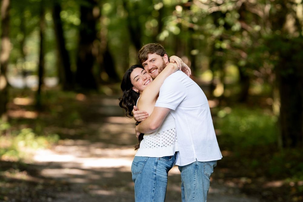 A couple embraces warmly on a sunny forest path, surrounded by lush green trees in a peaceful outdoor setting.