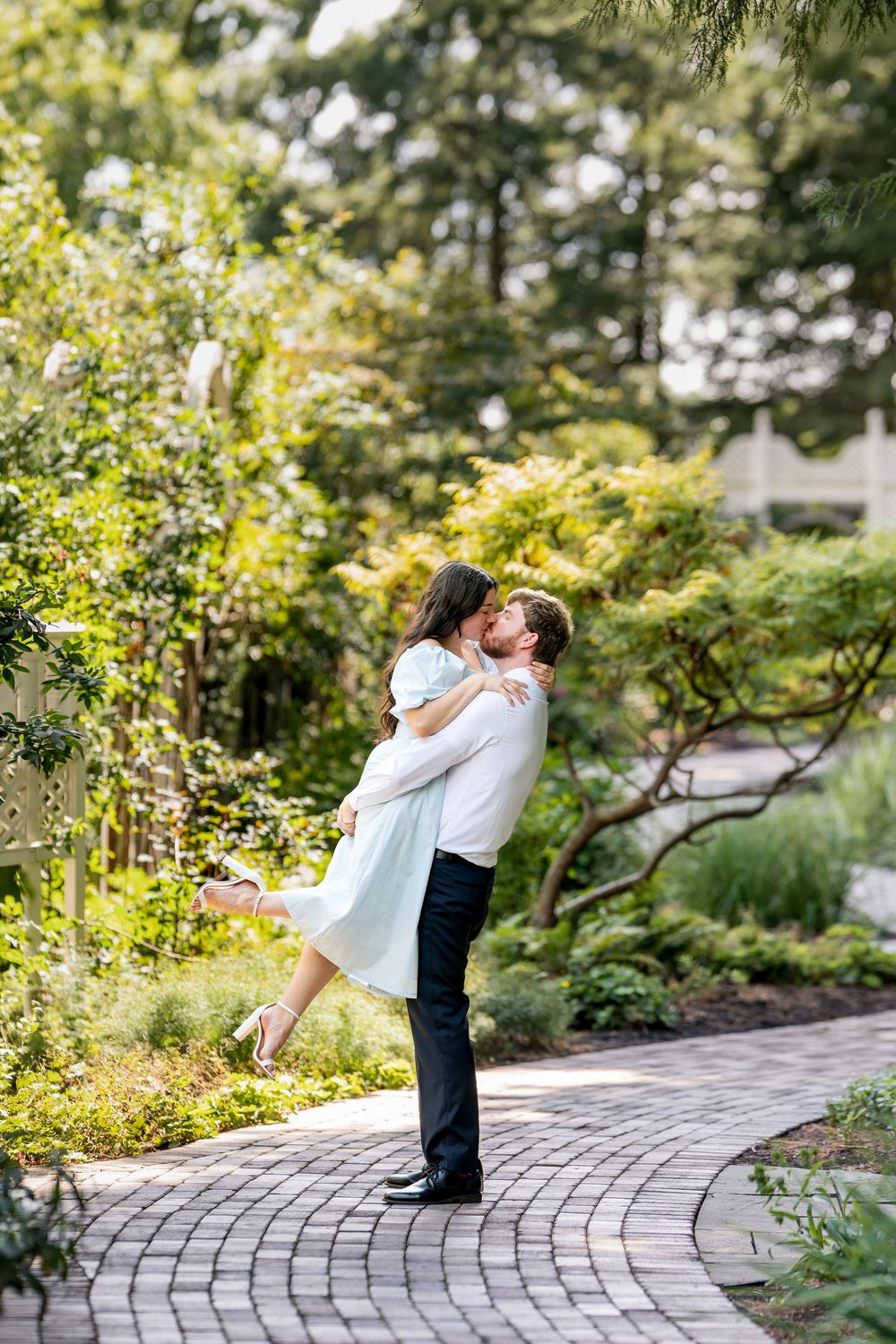 Couple sharing a romantic kiss as man lifts woman in a lush garden pathway.