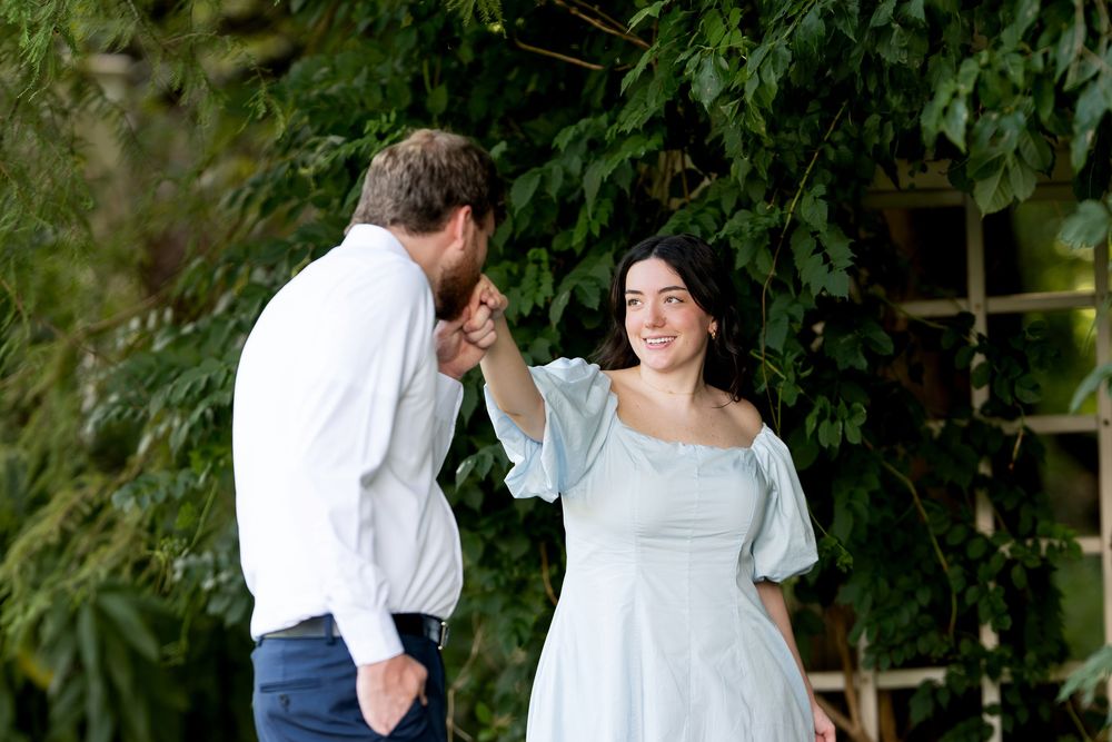A couple shares a romantic moment as the man kisses the woman's hand outdoors among lush green foliage.