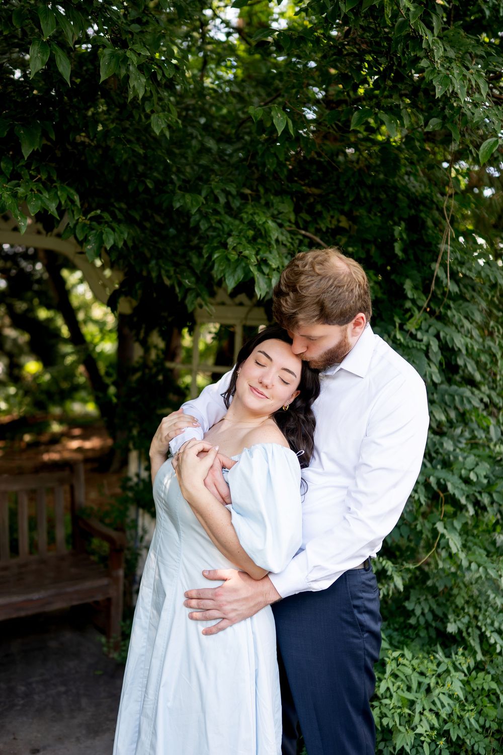 Man tenderly kissing woman's head while embracing her from behind under green garden arbor.