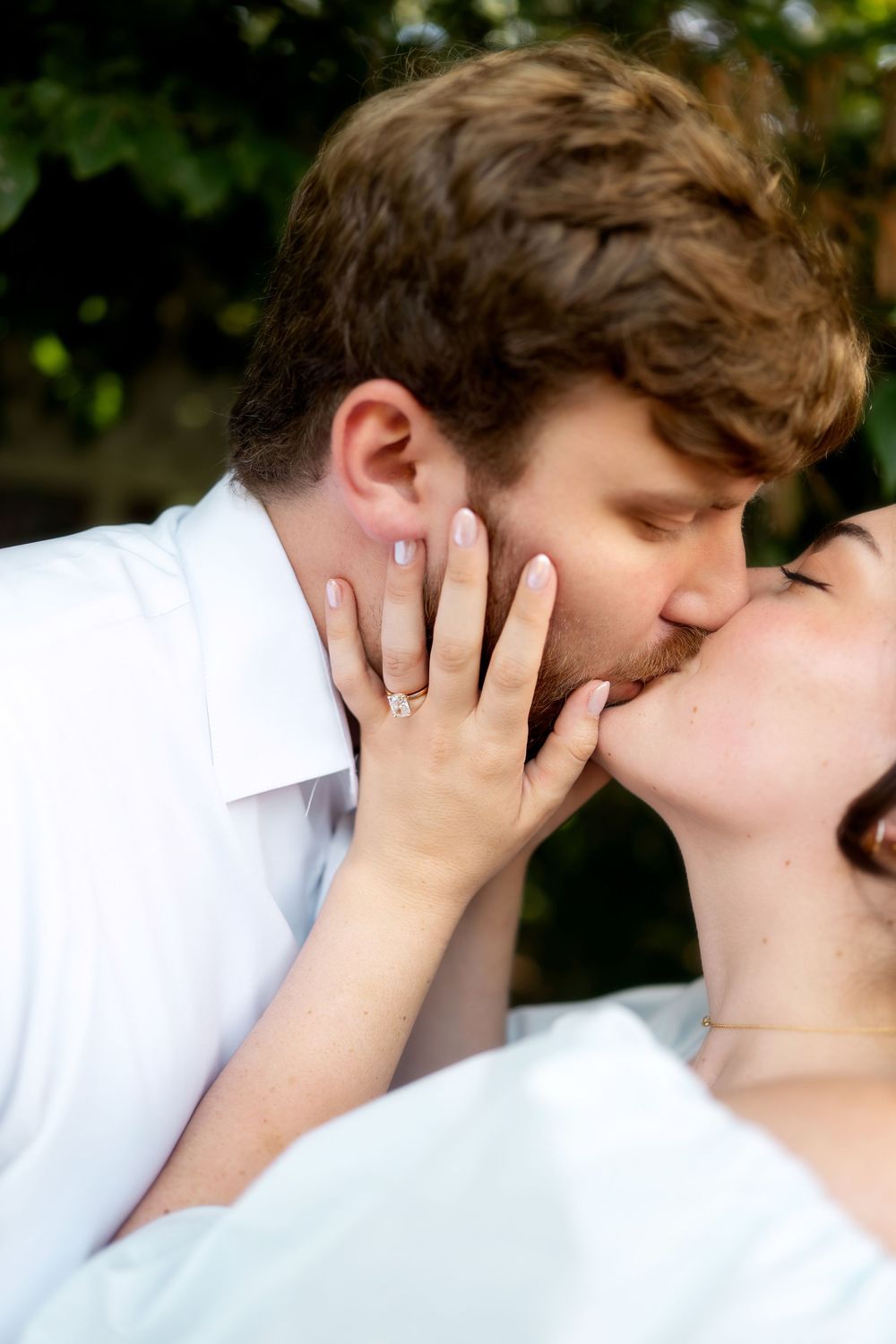 An engaged couple shares a tender kiss outdoors, with the woman's diamond engagement ring prominently displayed.