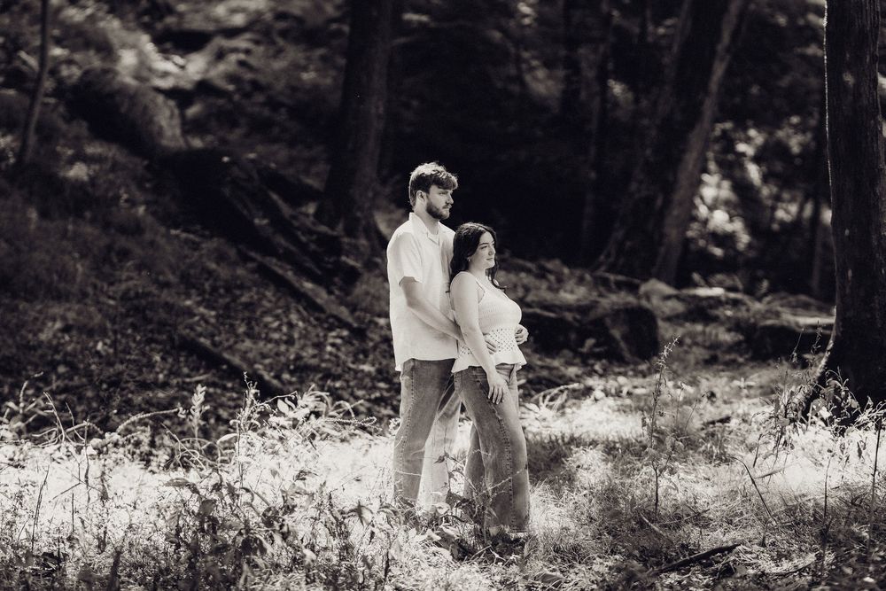 Black and white photo of a couple embracing in a lush forest setting, surrounded by trees and wildflowers.