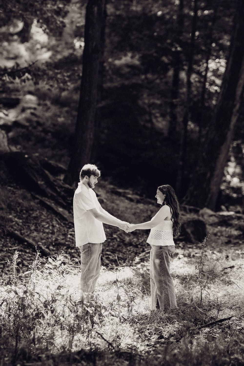 Black and white photo of couple holding hands in a sunlit forest clearing.