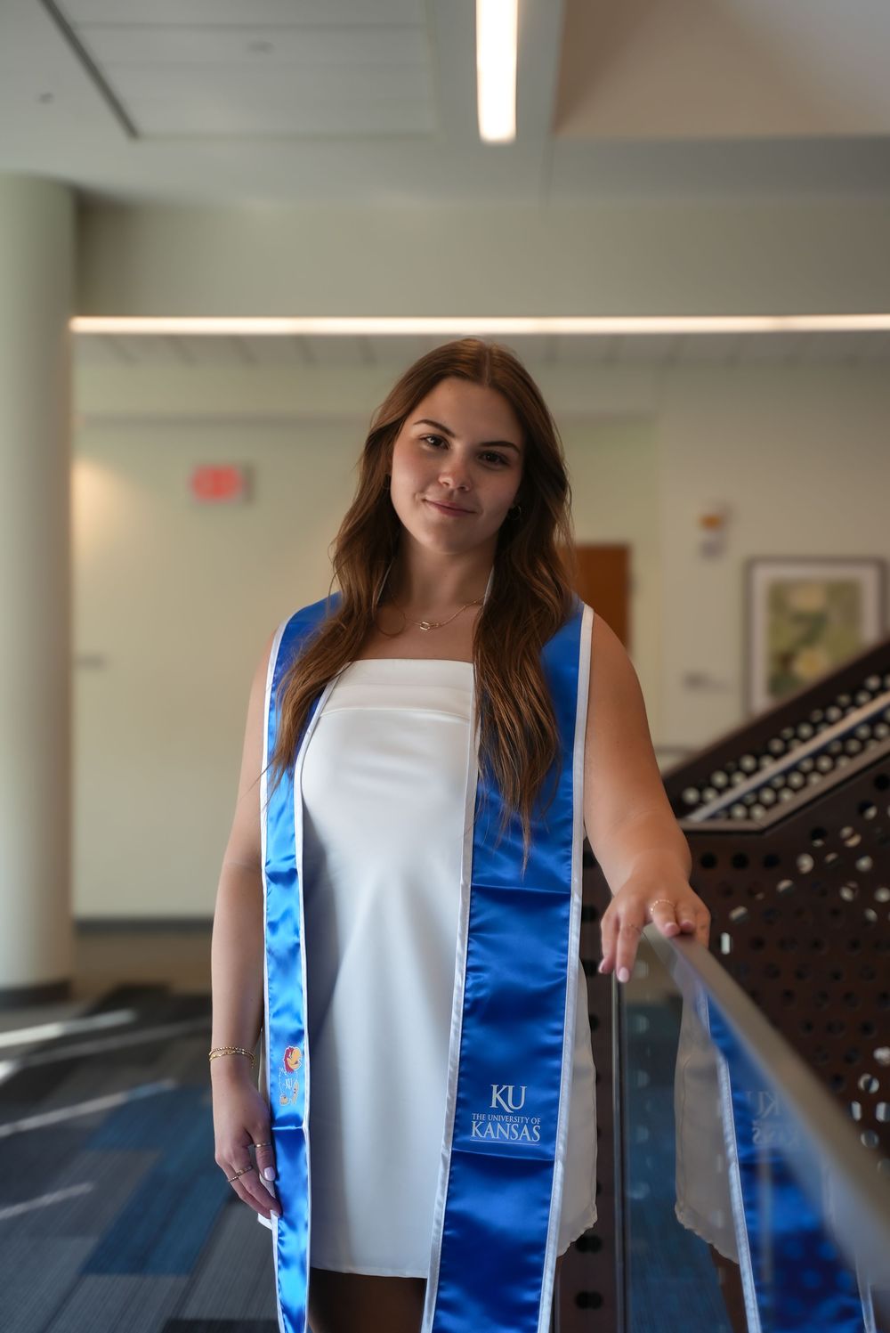 University of Kansas graduate in white dress wearing blue KU graduation stole, posing indoors near staircase railing.