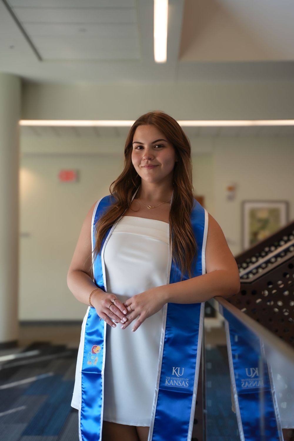 University of Kansas graduate smiling in white dress with blue KU graduation stole indoors.