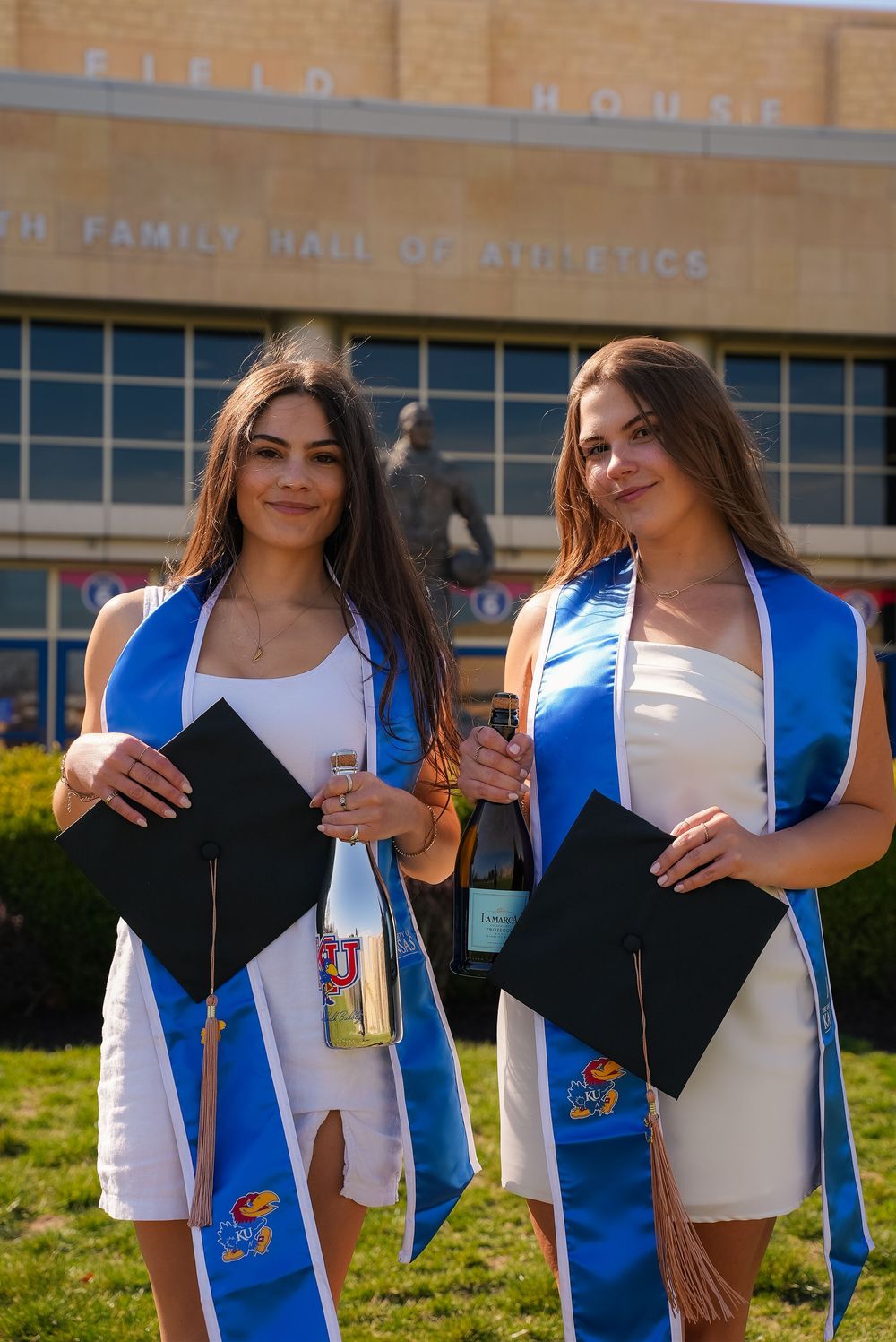 Two female graduates in white dresses with blue KU stoles holding caps and bottles outside Field House.